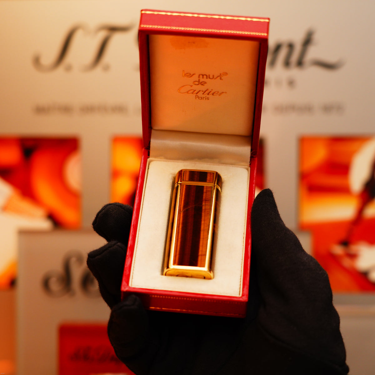 A gloved hand holds an open red box showing a Vintage 1970 Cartier Tiger's eye Solid 18k lighter (750 hallmark) from KirbyAllison.com, with "les must de Cartier Paris" inscribed inside.