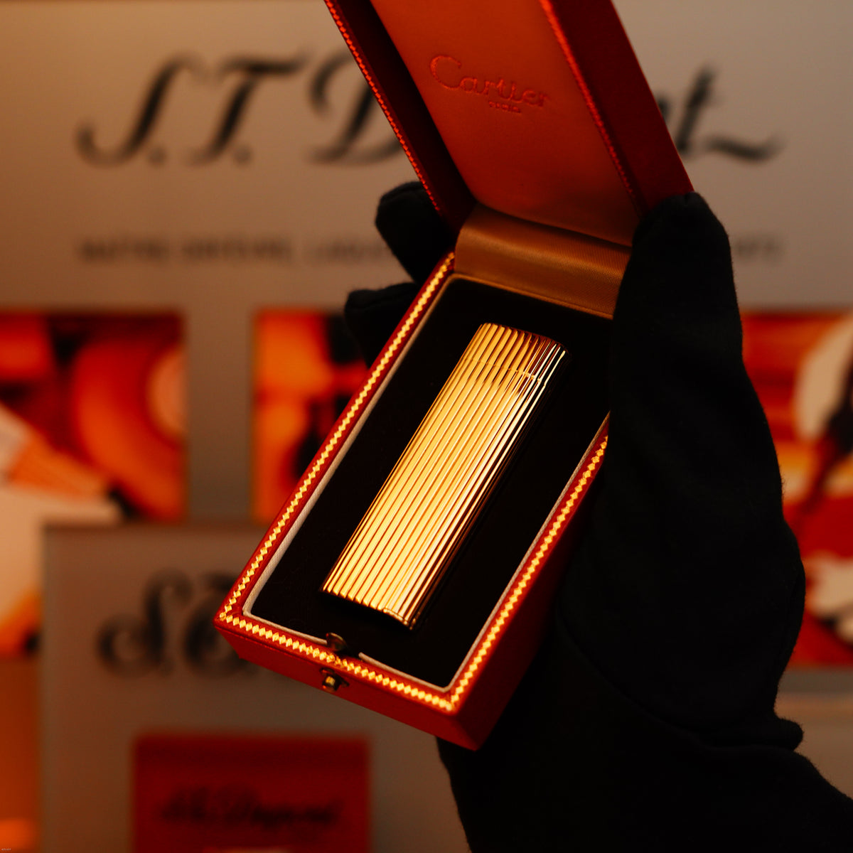 A gloved hand holds an open box displaying a Vintage 1970 Extremely Rare Cartier Solid 18k Gold Vertical Line Heavy Lighter from KirbyAllison.com, with a blurred S.T. Dupont display in the background.