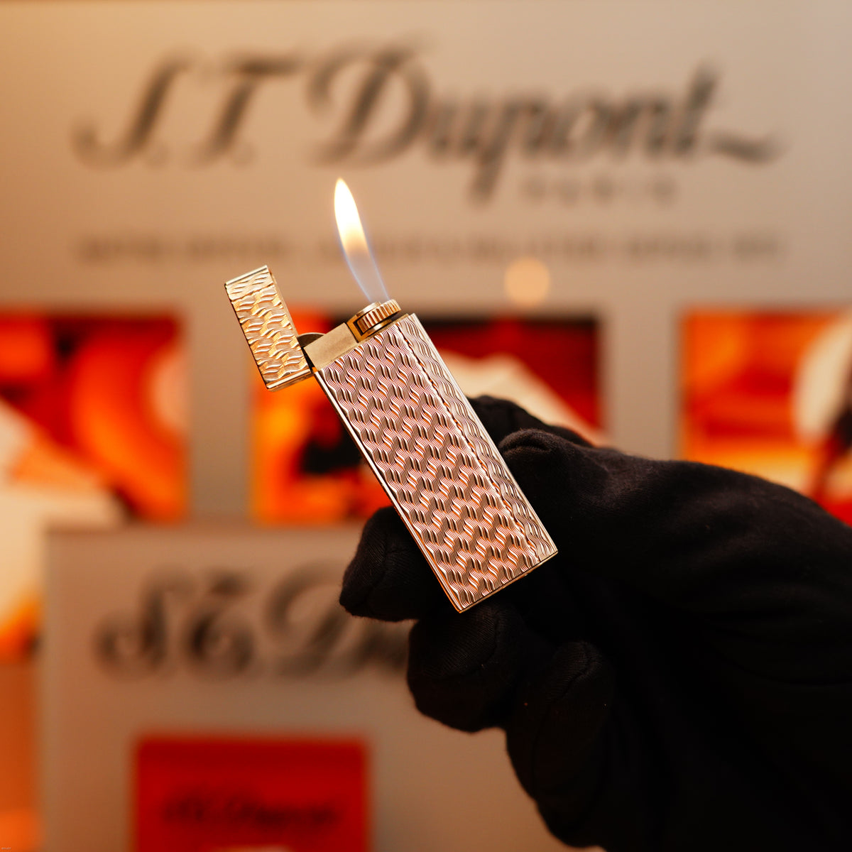 A gloved hand holds a lit Vintage 1970 Early Cartier Production Pentagone Solid 925 Heavy Sterling Silver Lighter by Cartier, its textured metal surface on display against a blurred branded background.