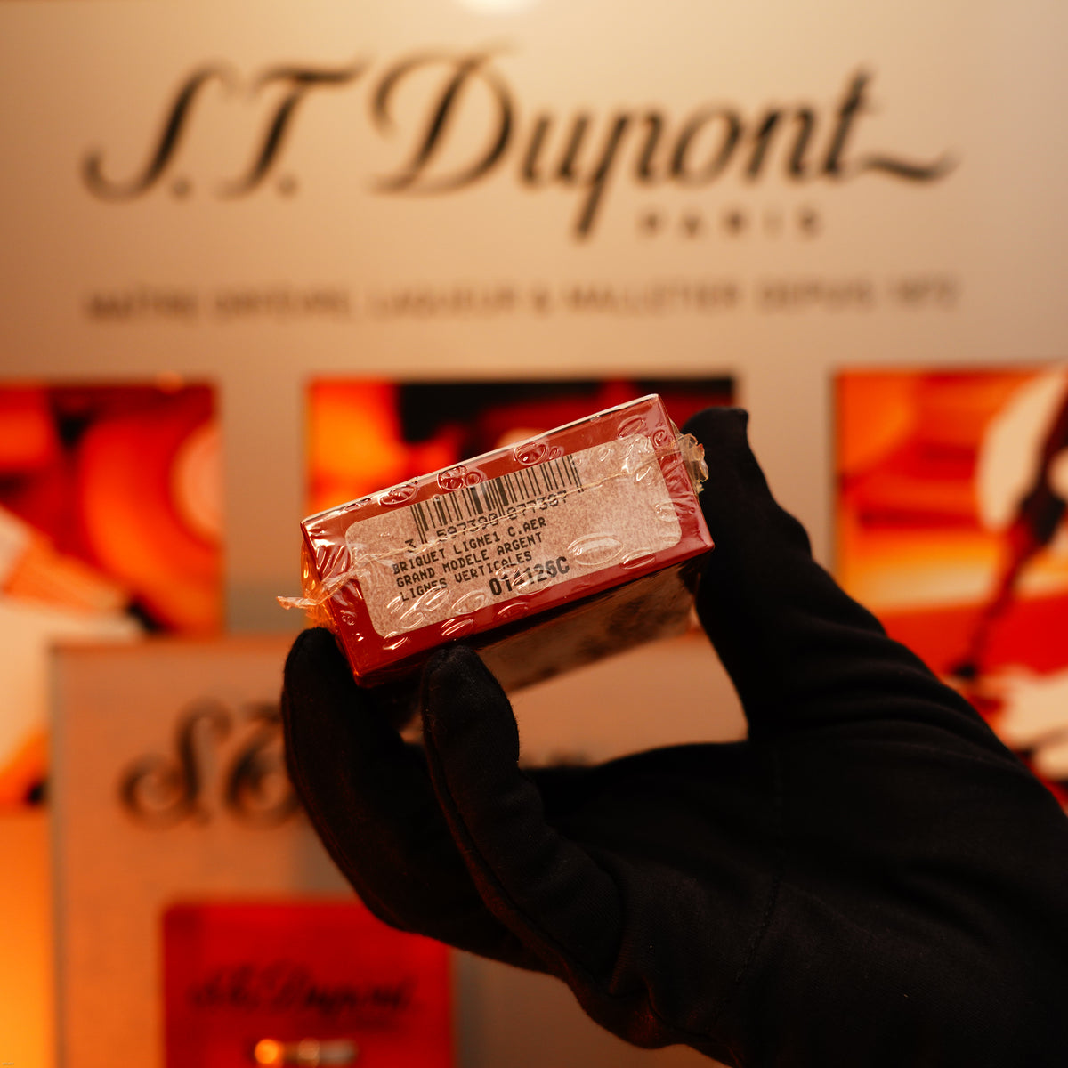 A gloved hand holds a red Lucy Lighter box with a barcode and label, showcasing quality guarantee, in front of a display sign for the Vintage 1990 St Dupont Heavy Silver iconic Vertical ligne rare Sealed Lighter.