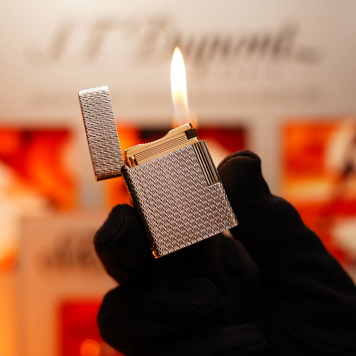 A gloved hand holds the Lucy Lighter Vintage 1970 Extremely Rare 1/1 De St Dupont Solid 925 Sterling Ligne 1 Guilloché Pattern Lighter with its lid open and flame burning—luxuriously crafted and backed by a 12-month warranty.