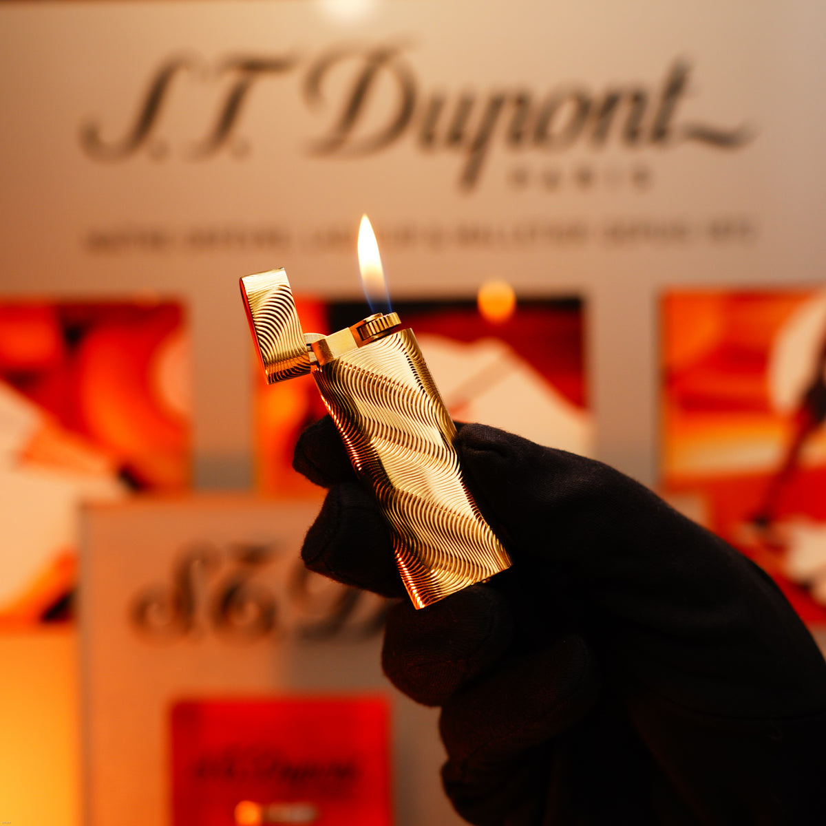 A gloved hand holds a Lucy Lighter Vintage 1970 Cartier, an extremely rare solid 18K gold lighter with heavy guilloché pattern and 750 hallmark, lid open and flame lit, against a blurred branded background.