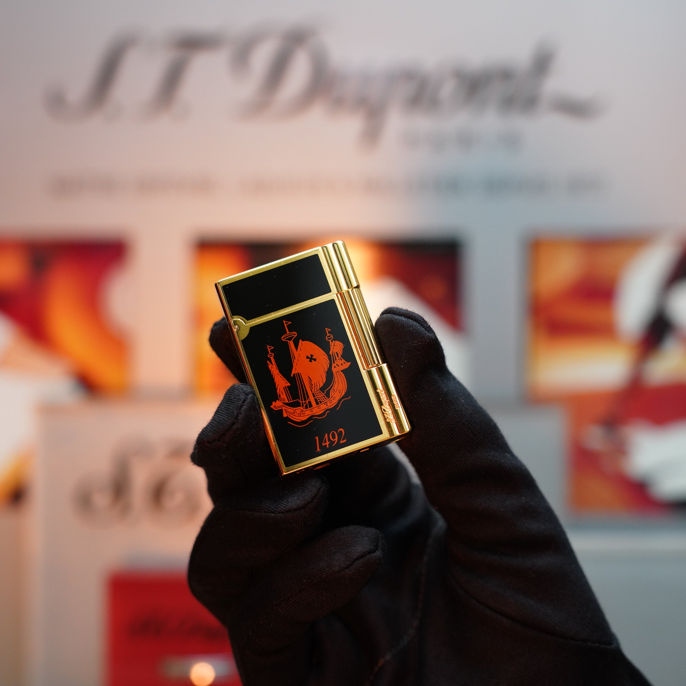A gloved hand holds the second limited edition ever made of the Vintage 1992 St Dupont Columbus Gatsby lighter, adorned in an elegant 24k gold finish with black detailing. Featuring the "1492" inscription and a ship design, the S.T. Dupont branding in the blurred background subtly evokes its timeless charm.