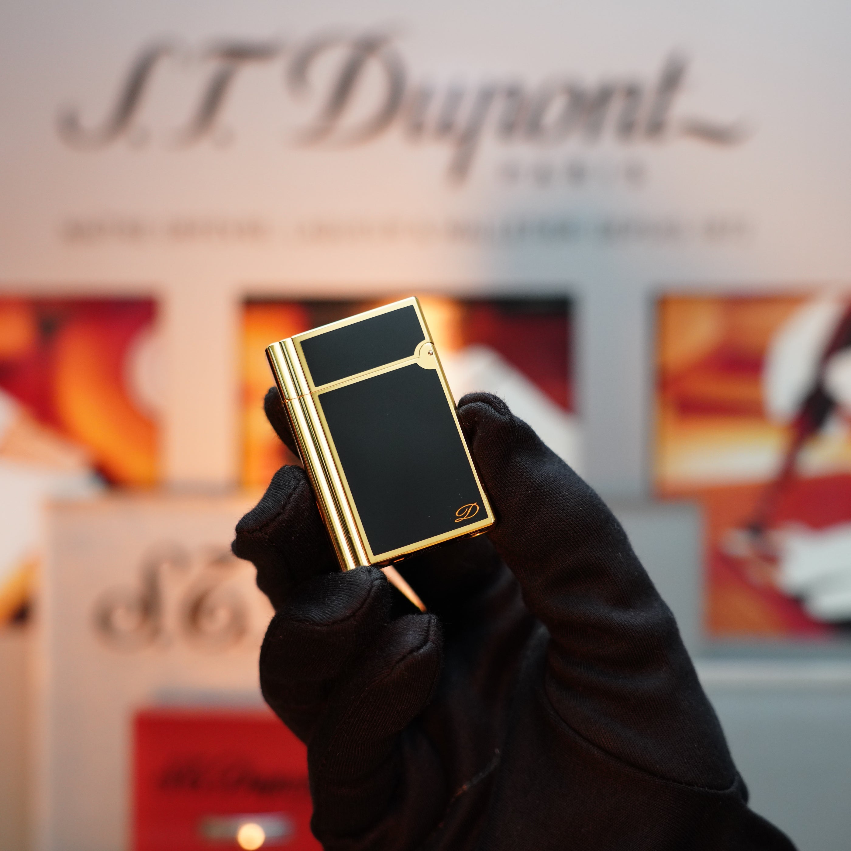 A gloved hand delicately holds a Vintage 1992 S.T. Dupont Important 24k Gold Finish Colombus Gatsby second Limited Edition ever made lighter, featuring a black and gold design, set against a blurred background showcasing the brand's logo and capturing the essence of timeless elegance.
