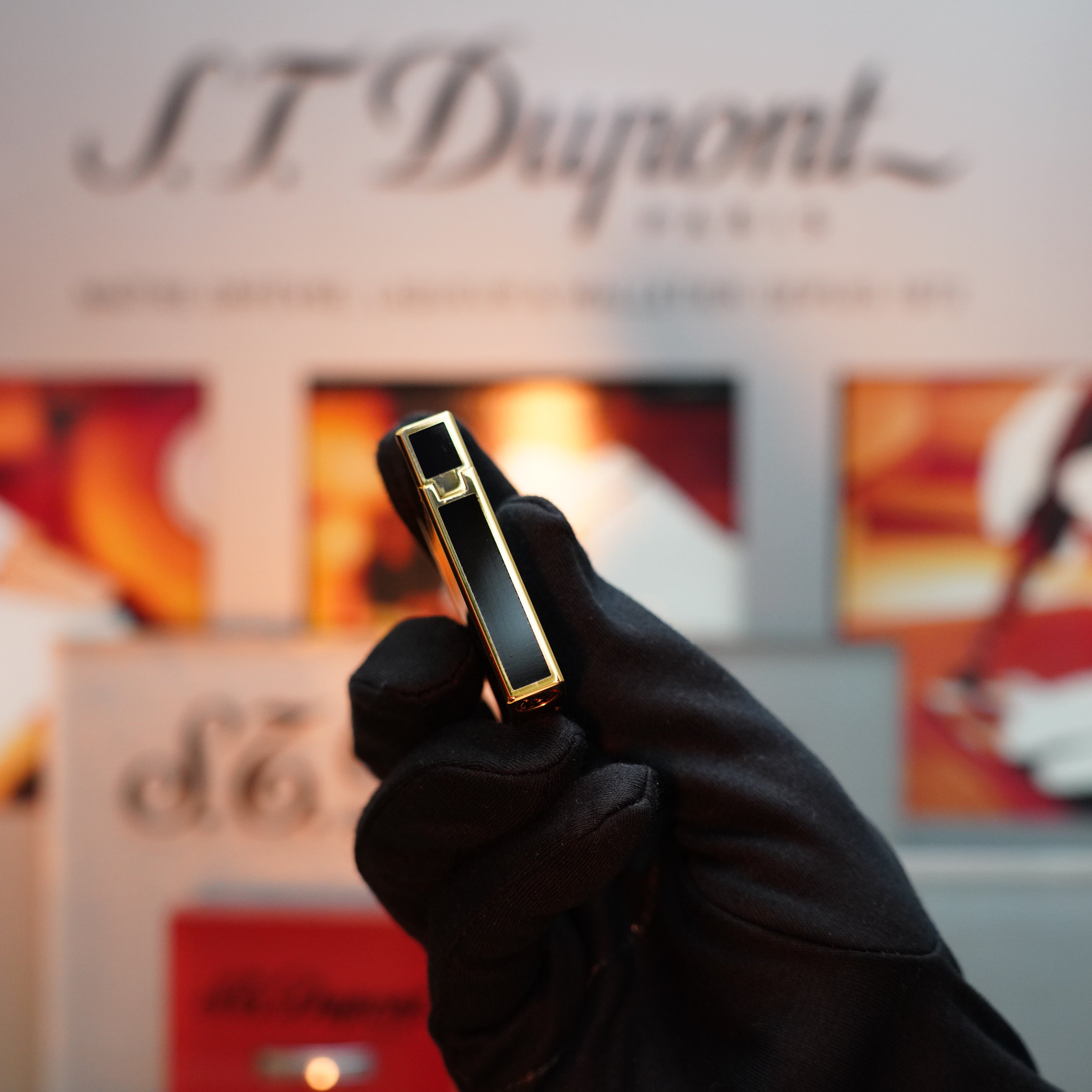 A person wearing a black glove holds a stylish, Vintage 1992 S.T. Dupont Colombus Gatsby lighter with an important 24k gold finish, which is the second Limited Edition ever made, against a background with a blurred brand logo.
