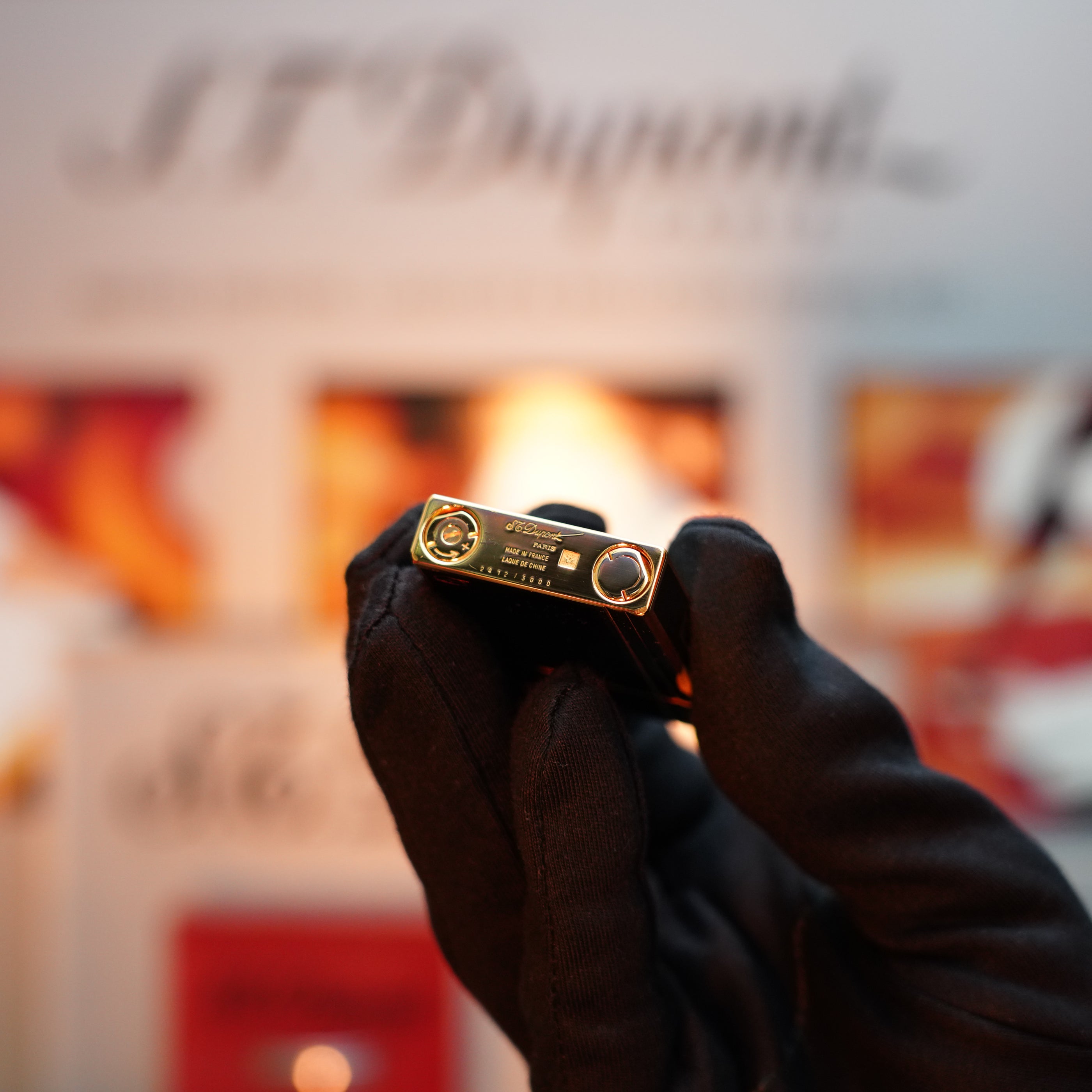 A gloved hand holds up a limited edition Vintage 1992 S.T. Dupont Colombus Gatsby lighter, featuring a 24k gold finish and metallic details, against a blurred background.