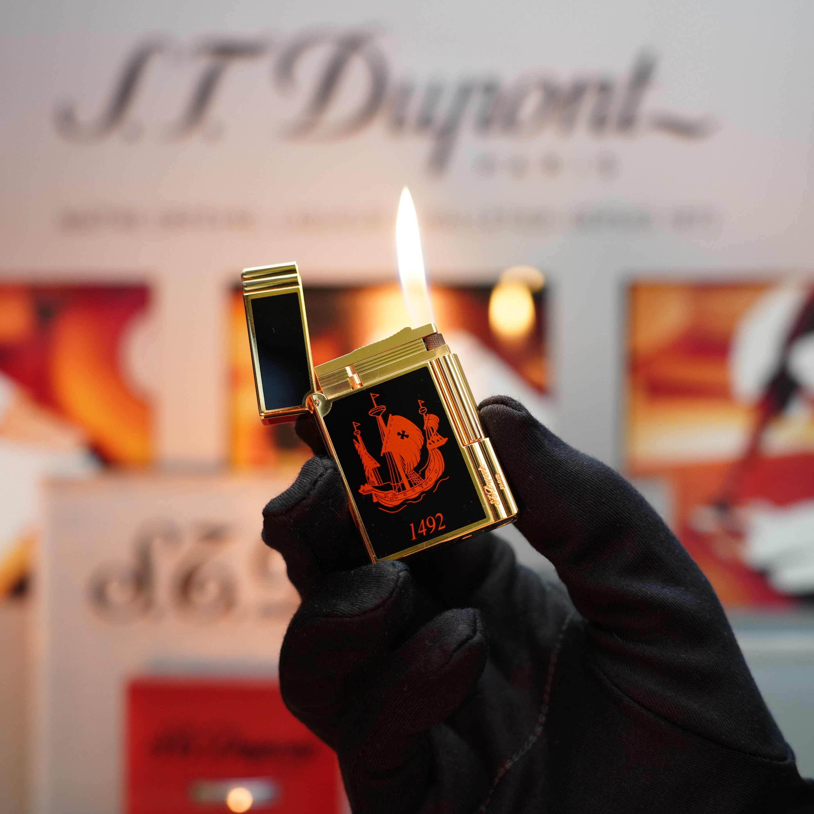 A gloved hand holds a Vintage 1992 S.T. Dupont Important 24k Gold Finish Colombus Gatsby second Limited Edition ever made lighter with a lit flame, highlighting its black and gold design and the "1492" inscription, which echoes the elegance of vintage craftsmanship.