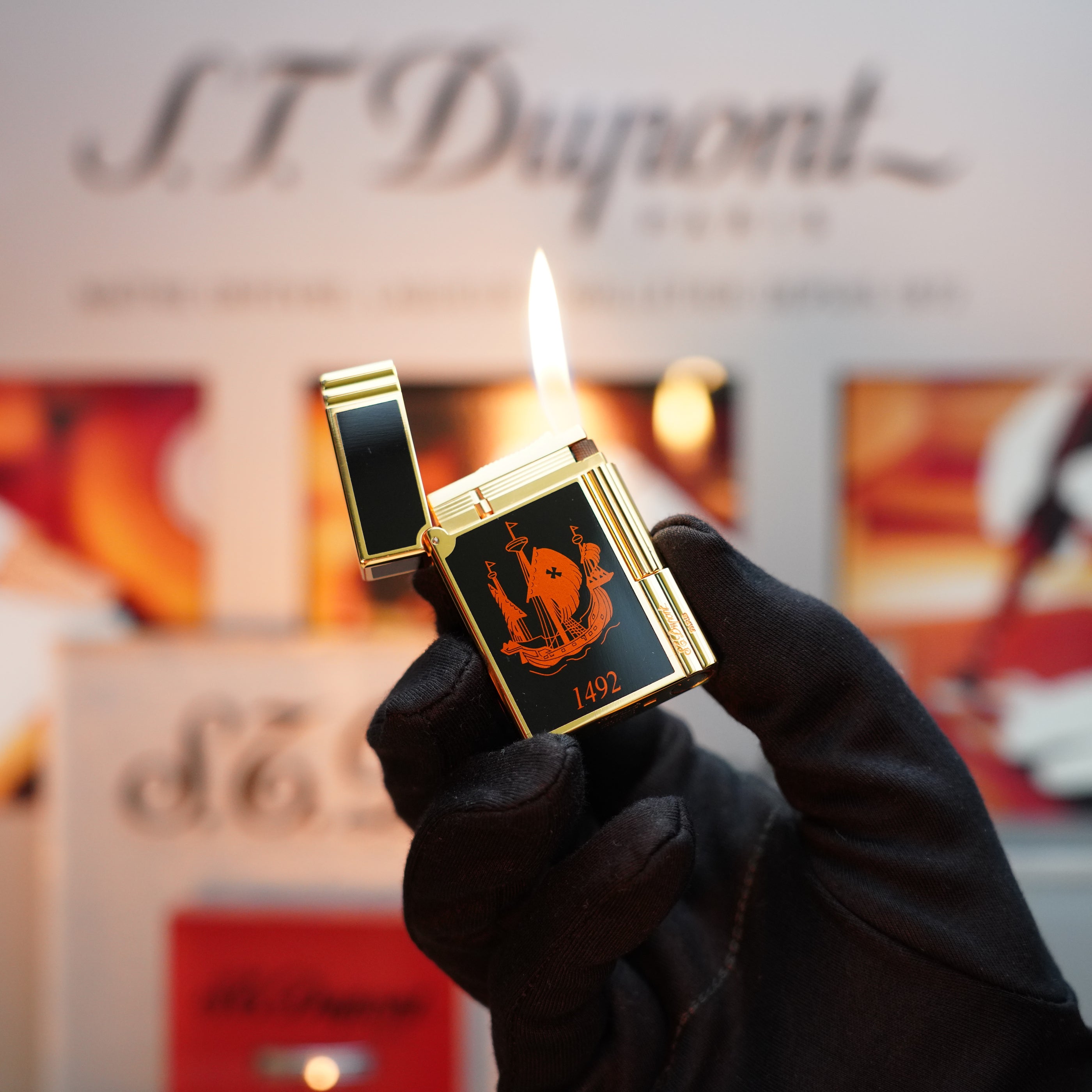 In a gloved hand, a person holds an open Vintage 1992 S.T. Dupont Important 24k Gold Finish Columbus Gatsby Second Limited Edition Lighter, elegantly showcasing its orange flame against the backdrop where the prestigious brand name "S.T. Dupont" is prominently displayed, emphasizing its exclusive allure.