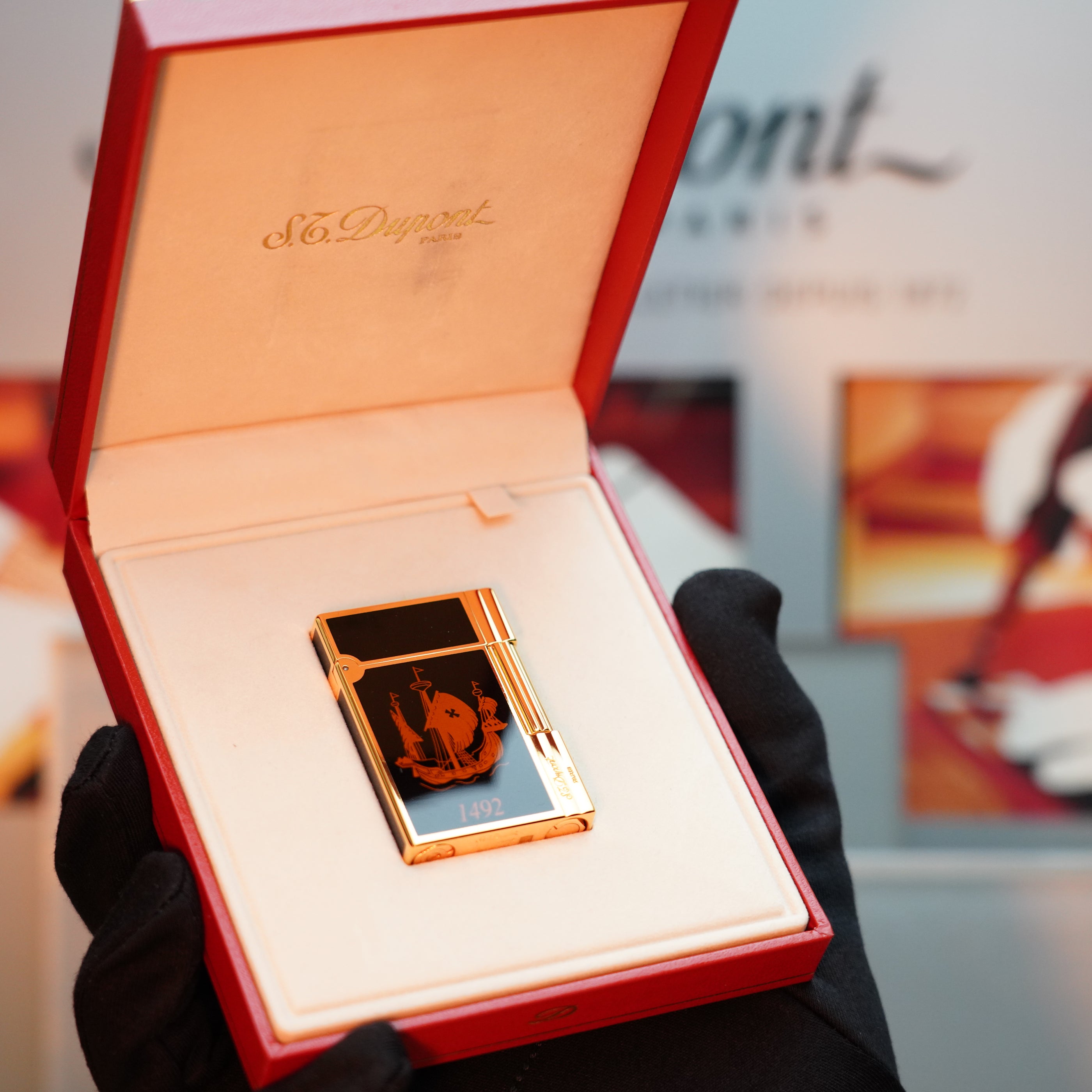 A hand in a glove carefully holds an open red box, revealing a Vintage 1992 S.T. Dupont Important 24k Gold Finish Colombus Gatsby second Limited Edition ever made Lighter with an elegant gold and black engraved design inside.