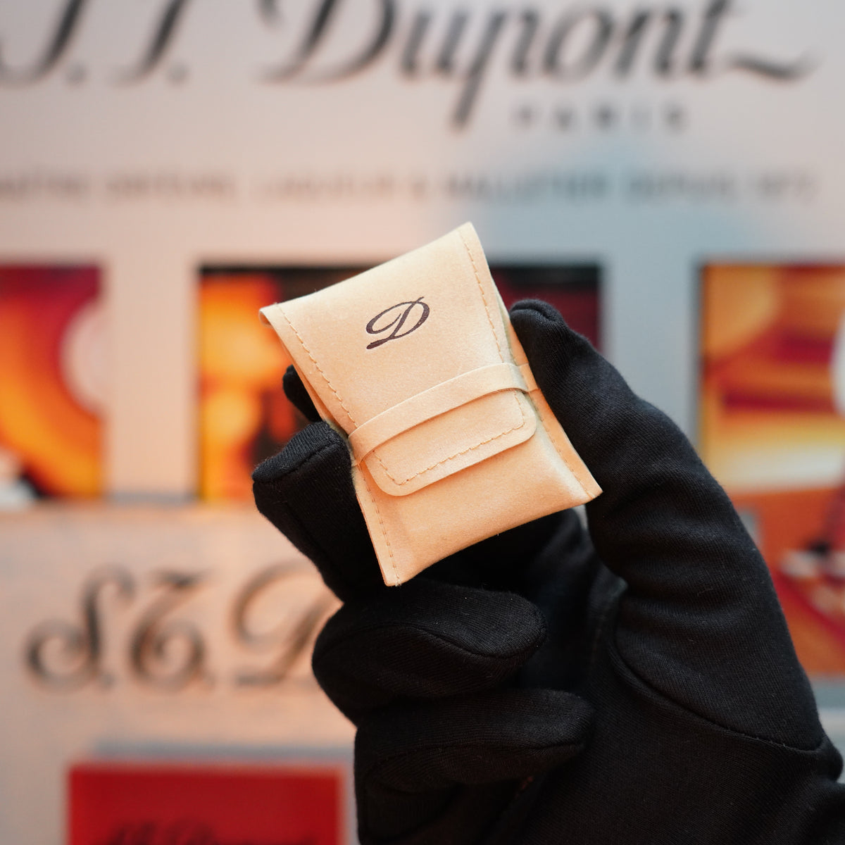 A gloved hand holds a small beige pouch with the letter "D" on it, in front of a blurred S.T. Dupont display, evoking the elegance of the Vintage 1990 St Dupont Iconic 24k Gold Finish D Ligne Full Hand Applied Lacquer with Lacquer Wheel Lighter.
