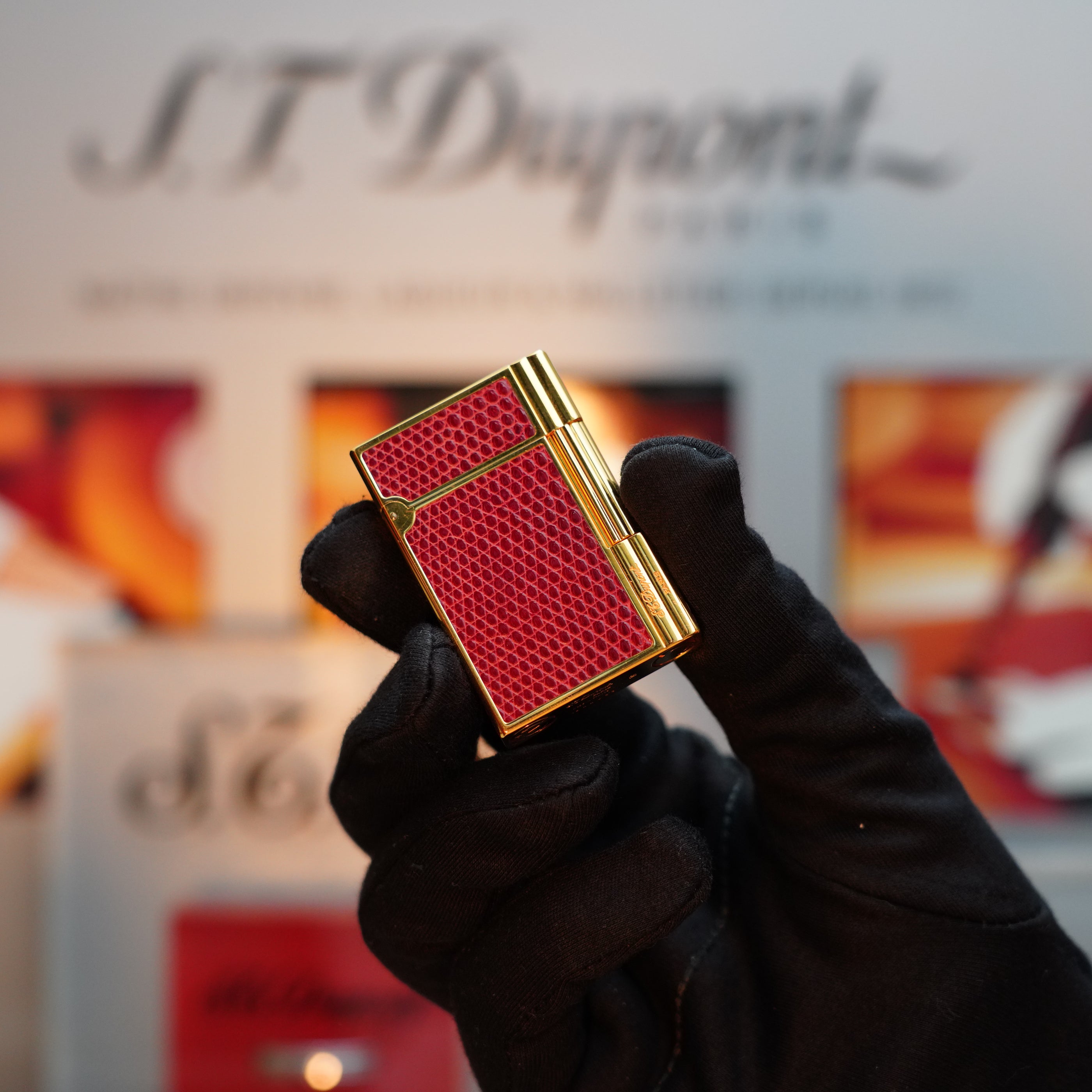 A gloved hand holds the vintage 1980 S.T. Dupont Gatsby Gainerie Lezard Red Bordeaux lighter, featuring an extremely rare authentic leather design and a 24k Gold finish, set against a blurred S.T. Dupont display background.