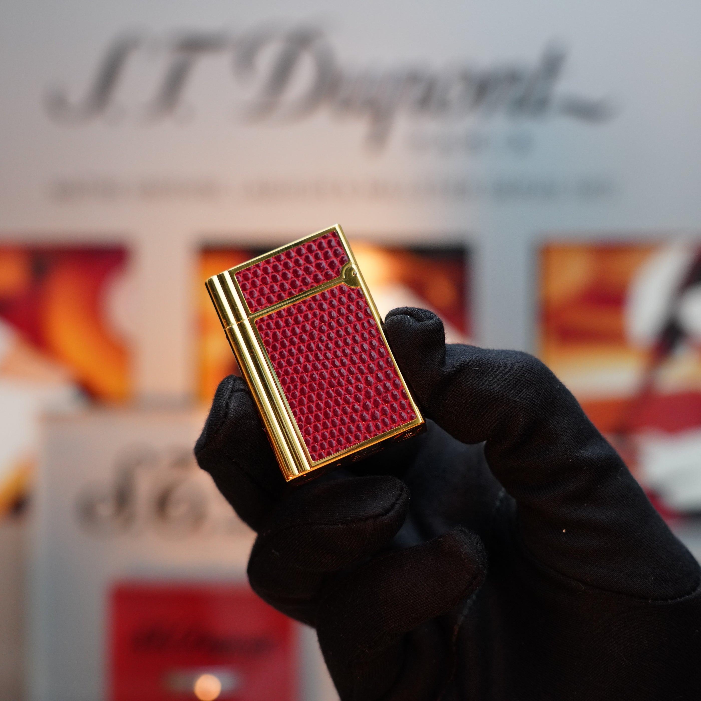 A gloved hand holds an extremely rare Vintage 1980 S.T. Dupont Gatsby Gainerie Lezard lighter, featuring a 24k gold finish and an authentic red Bordeaux leather textured design, in front of a blurred background with the text "S.T. Dupont.