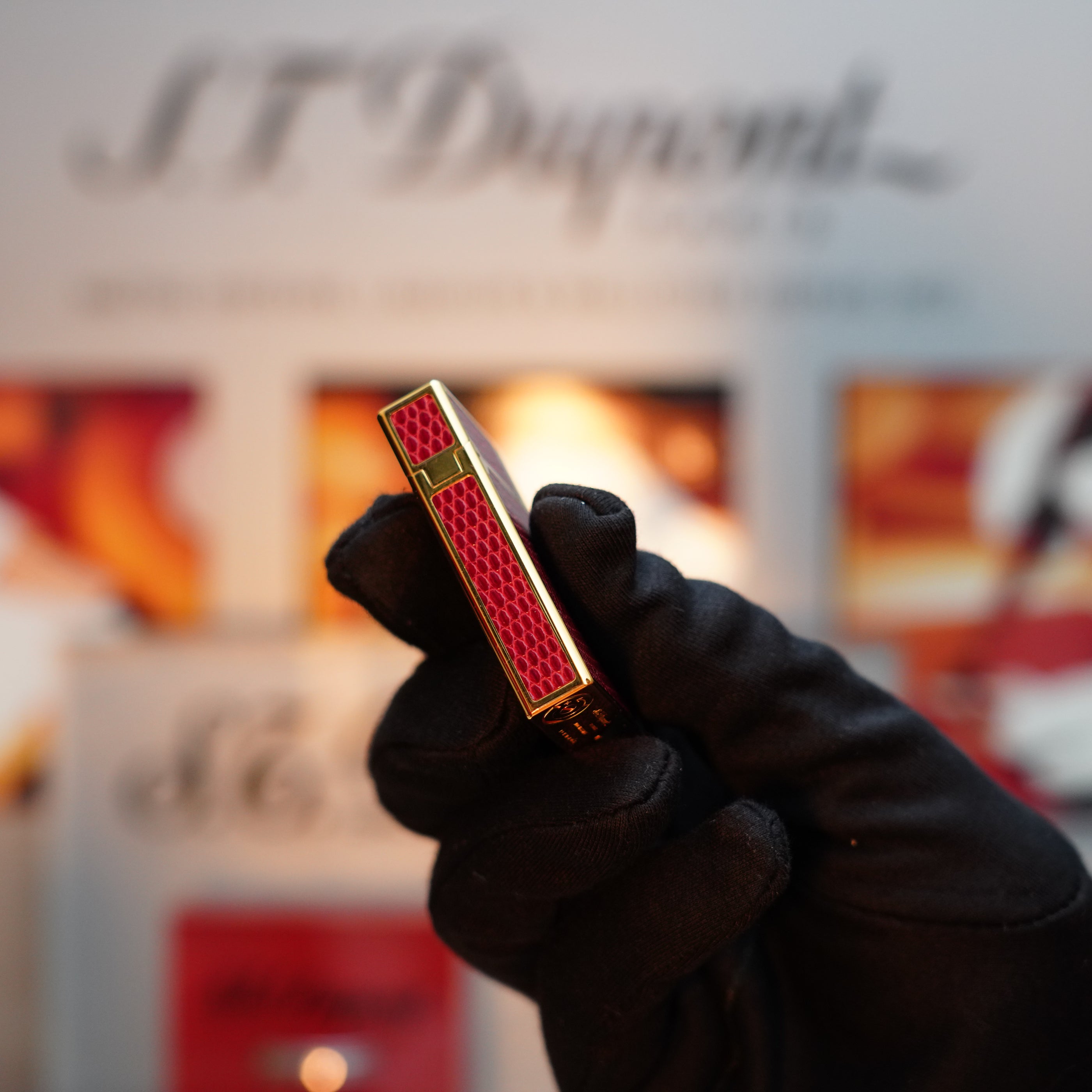 A gloved hand holds a Vintage 1980 S.T. Dupont Gatsby Gainerie Lezard Red Bordeaux lighter, featuring an authentic leather design with a 24k gold finish, set against a blurred backdrop.