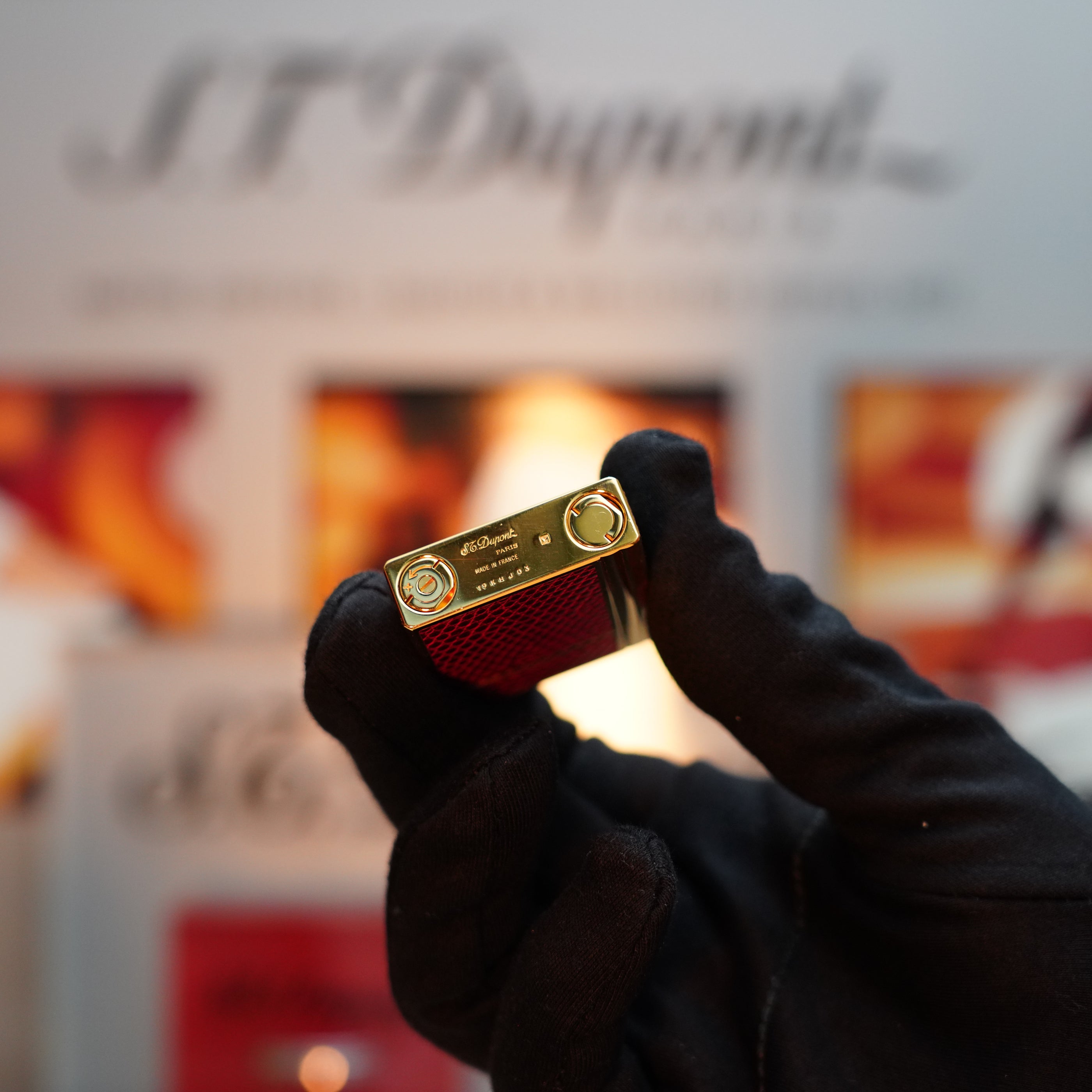 A black-gloved hand grasps a rare S.T. Dupont Vintage 1980 Gatsby lighter in red Bordeaux, featuring an exquisite 24k gold finish and authentic Lezard leather, set against a blurred backdrop.