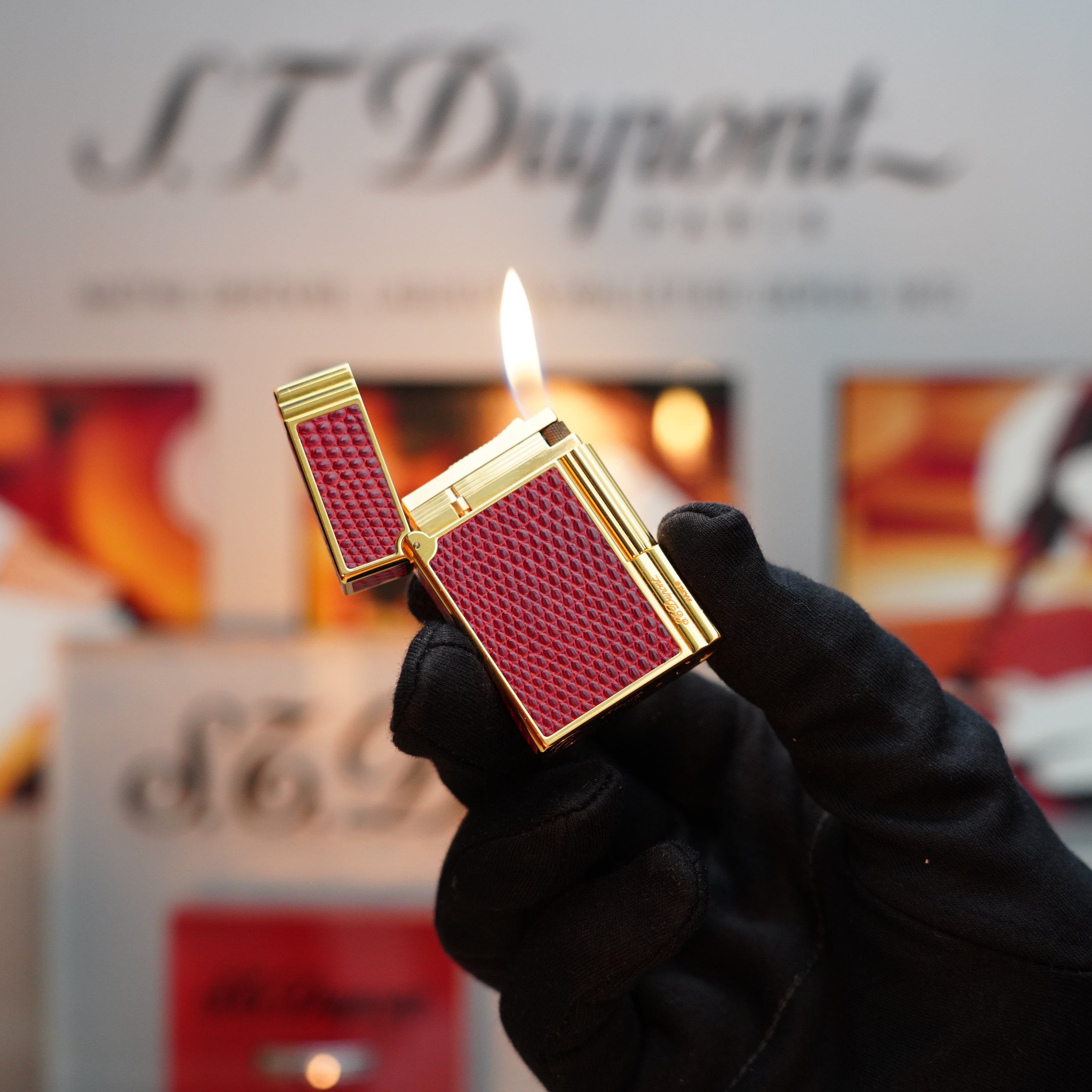A person wearing black gloves holds a lit S.T. Dupont Vintage 1980 Lighter with a 24k gold finish against a blurred background with text.