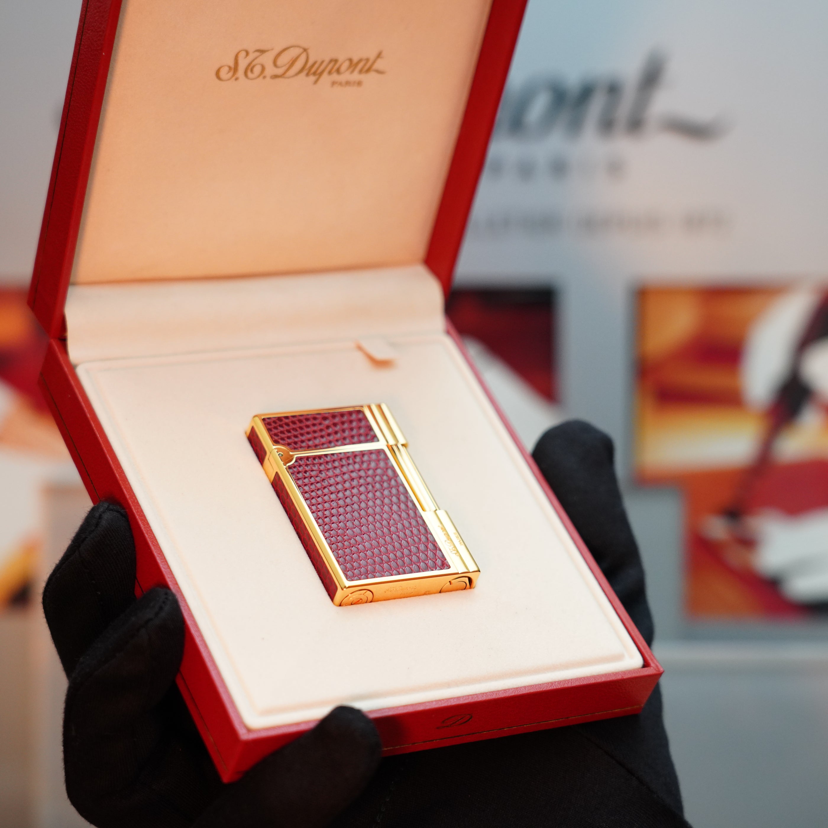 A gloved hand holds an open red box containing a Vintage 1980 S.T. Dupont Gatsby Lezard Red Bordeaux leather lighter, featuring an exquisite 24k gold finish.