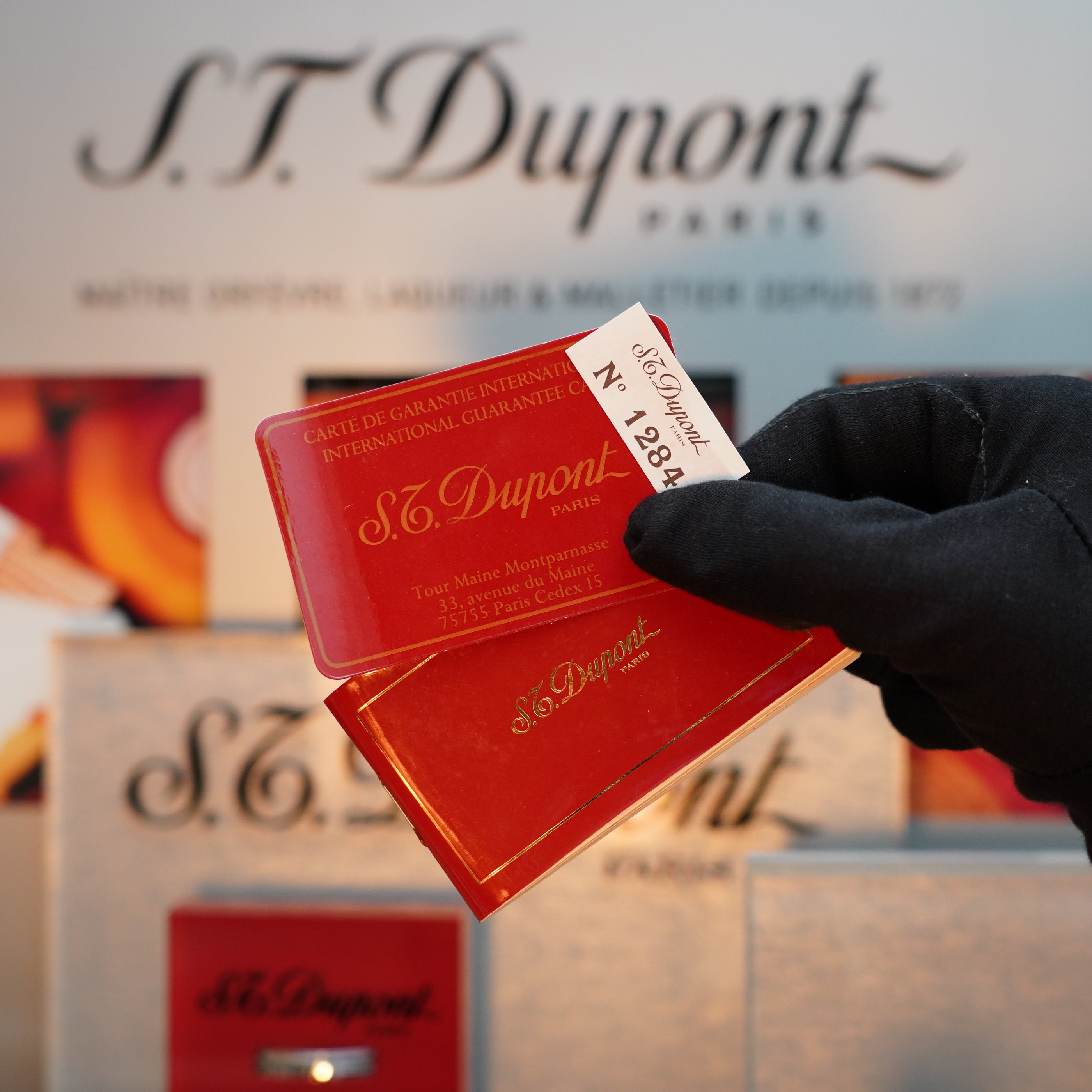 A gloved hand holds S.T. Dupont guarantee cards against a backdrop of S.T. Dupont branded boxes, echoing the sophisticated elegance of their Vintage 1980 24k Gold finish Gatsby Gainerie Lezard Red Bordeaux leather lighter collection.