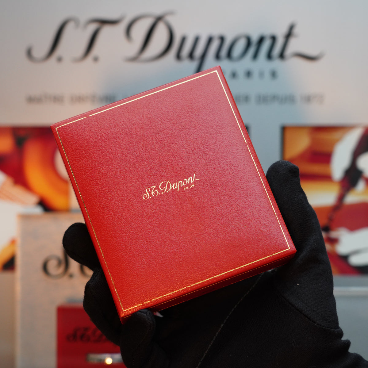 A gloved hand holds a red box adorned with the "S.T. Dupont Paris" text in gold, hinting at the exquisite craftsmanship of the Vintage 1980 S.T. Dupont 24k Gold finish Gatsby Gainerie Lezard Red Bordeaux Leather Extremely Rare Lighter inside, against a blurred background featuring the same iconic brand name.