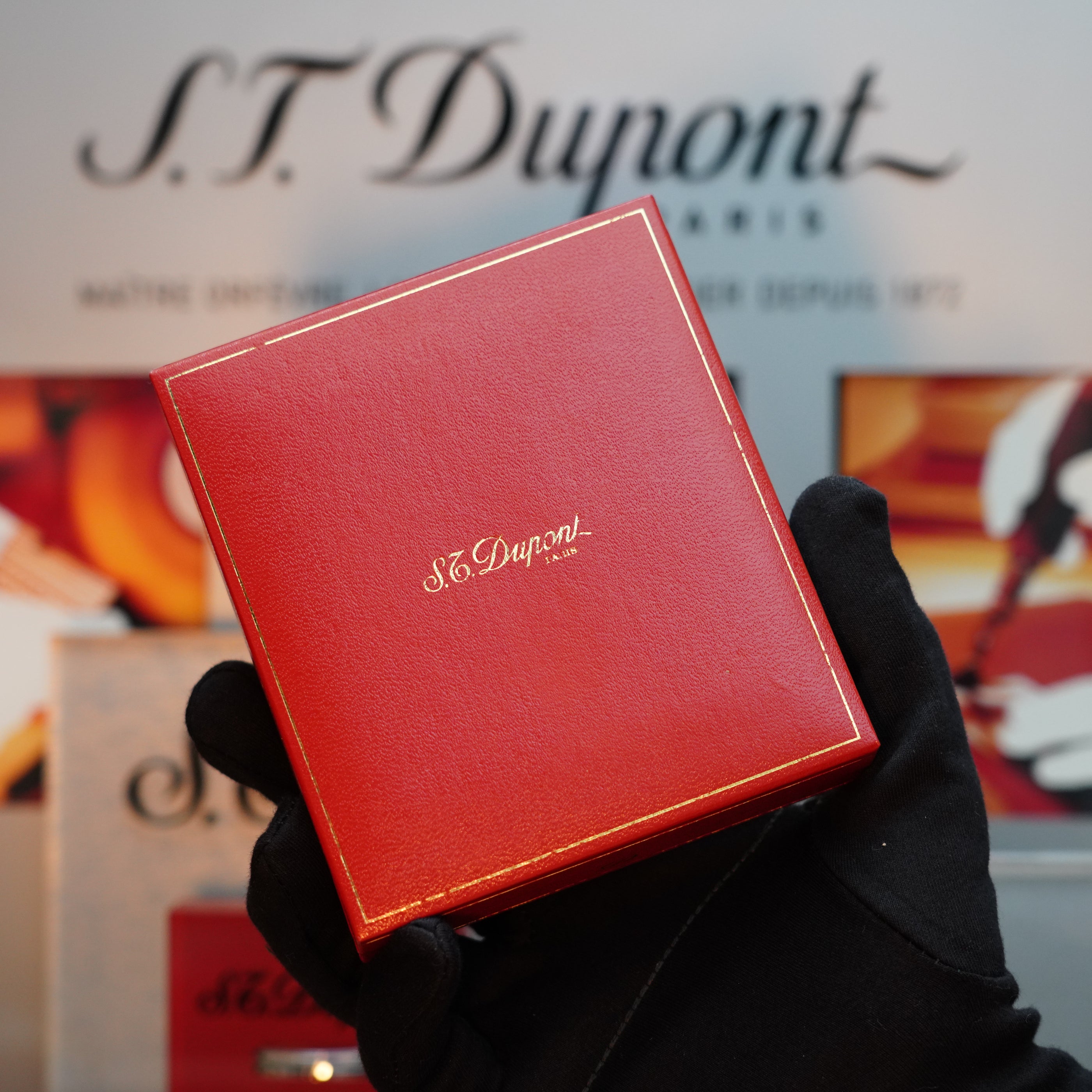 A gloved hand holds a red box adorned with the "S.T. Dupont Paris" text in gold, hinting at the exquisite craftsmanship of the Vintage 1980 S.T. Dupont 24k Gold finish Gatsby Gainerie Lezard Red Bordeaux Leather Extremely Rare Lighter inside, against a blurred background featuring the same iconic brand name.