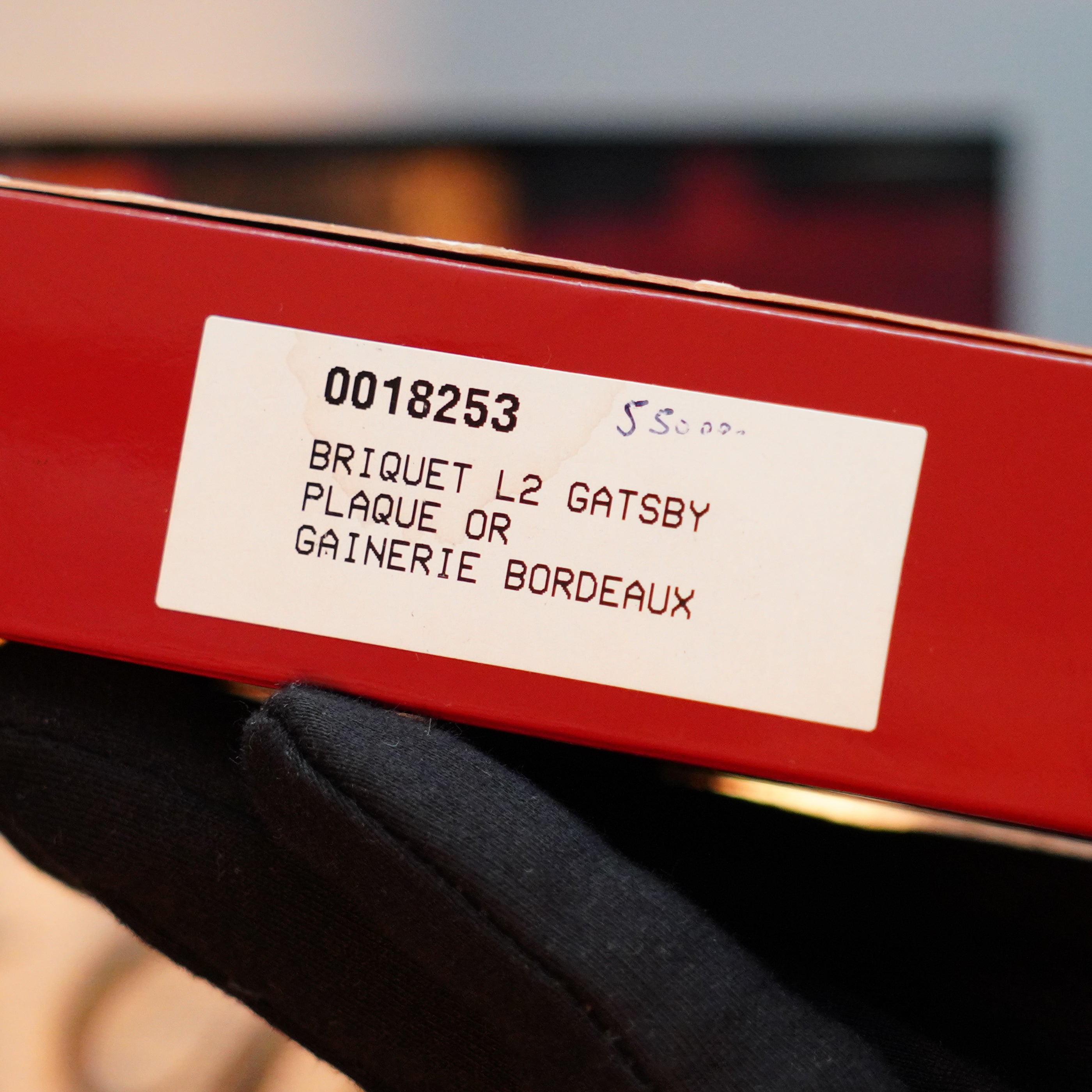 A close-up of a hand in a black glove holding a red box with a white label. The label reads "0008253 BRIQUET L2 GATSBY PLAQUE OR GAINERIE BORDEAUX," hinting at the exquisite 24k gold finish of the Vintage 1980 S.T. Dupont Gatsby Gainerie Lezard Red Bordeaux Authentic Leather Extremely Rare Lighter.