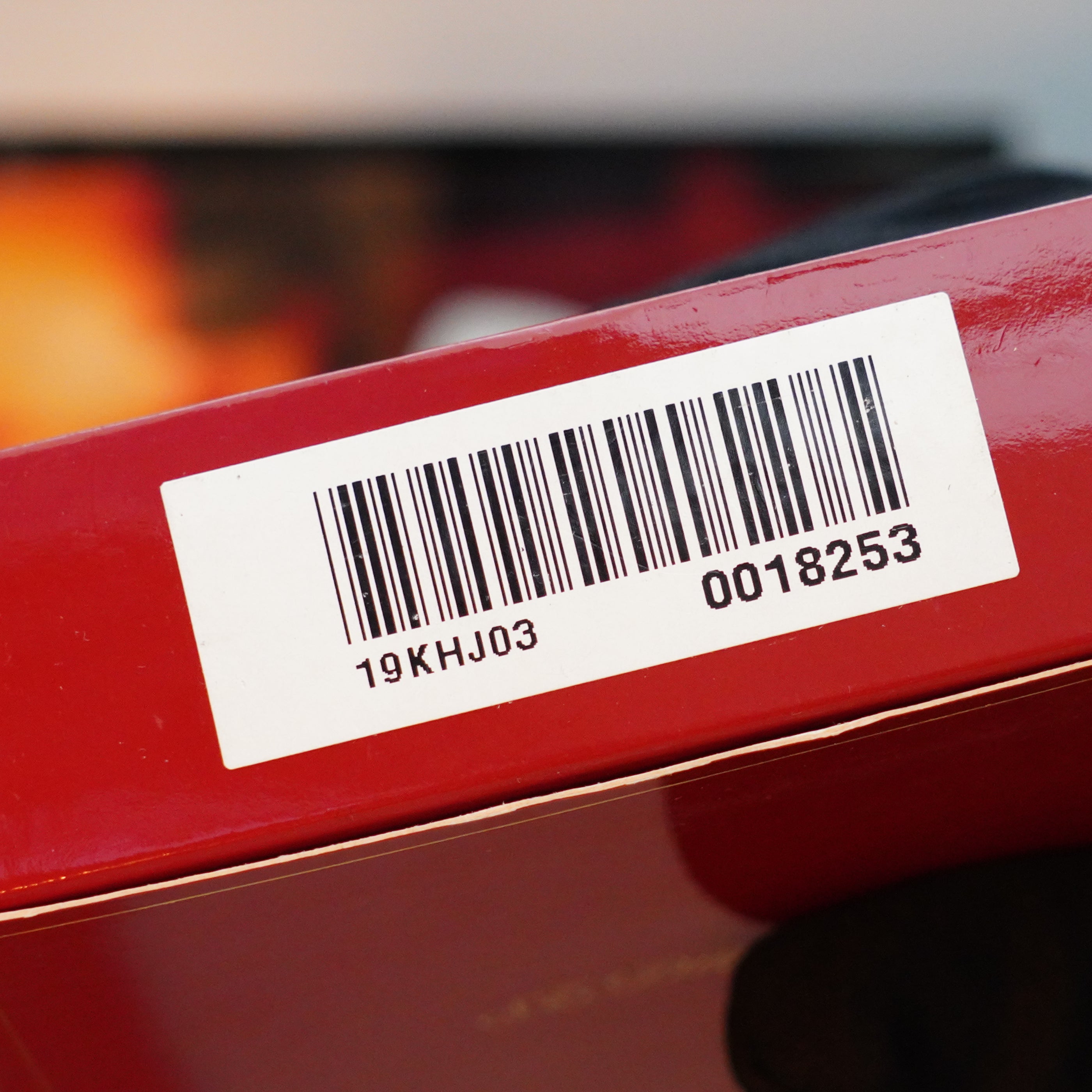 Close-up of a barcode sticker on a Vintage 1980 S.T. Dupont 24k Gold finish Gatsby Gainerie Lezard Red Bordeaux Authentic leather extremely Rare Lighter, resting on a red surface, displaying the numbers "19KHJ03" and "0018253".