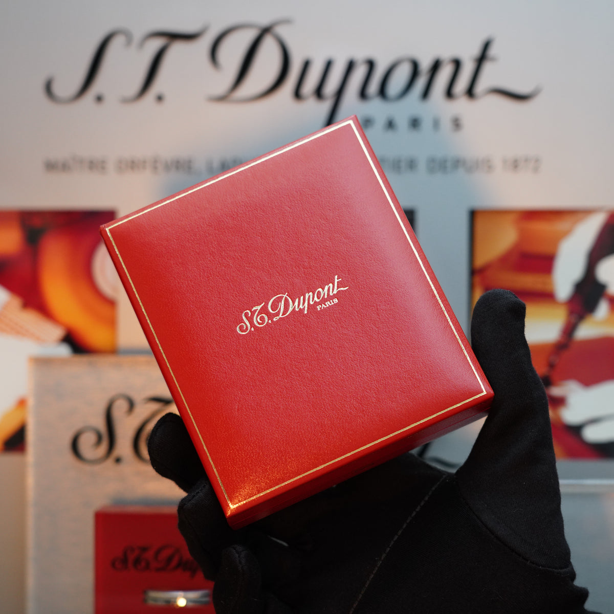 A gloved hand holds a red S.T. Dupont Paris box, reflecting the elegance of the Vintage 1980 St Dupont 24k Gold finish Gatsby Gainerie Lezard Brown Authentic leather extremely Rare Lighter against a branded background.