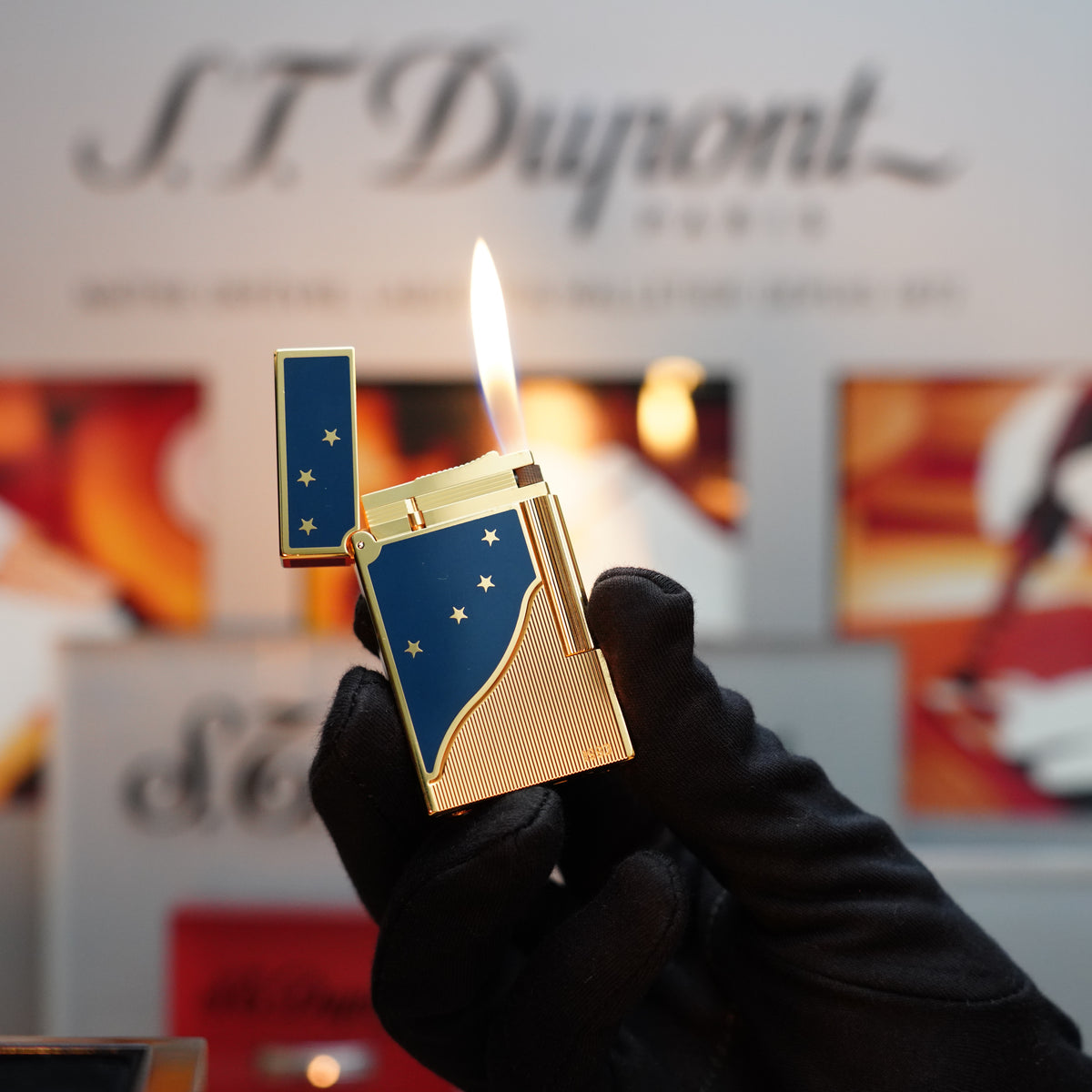A person wearing a black glove holds a Vintage 1993 S.T. Dupont Europe Rare 24k Gold Finish Blue Lacquer Limited Edition Ligne 2 Lighter with a lit flame, highlighting its exquisite design. The background displays blurred text and images.