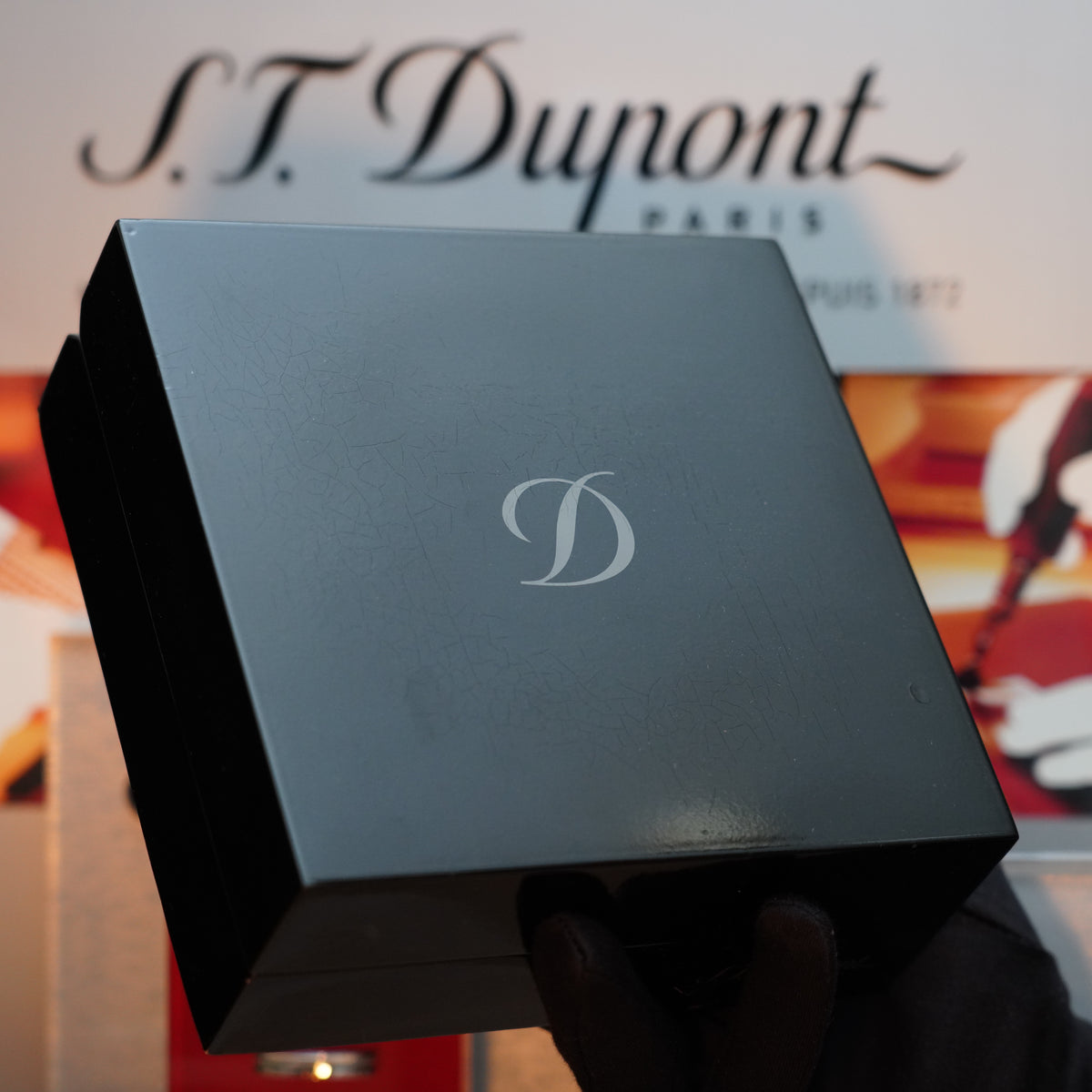 A gloved hand holds a special edition black box embossed with a silver "D" in front of an S.T. Dupont Paris display, evoking the vintage charm of a 1993 S.T. Dupont Ligne 2 lighter, renowned for its rare 24k gold finish and blue lacquer design.