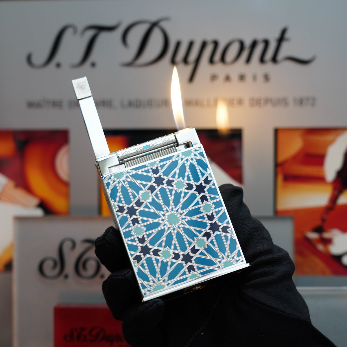 A gloved hand holds a lit, patterned S.T. Dupont Vintage 2003 Andalusia Limited Edition Blue Lacquer Table Lighter—one of only 300 made—against a blurred background showcasing the iconic brand's signage.
