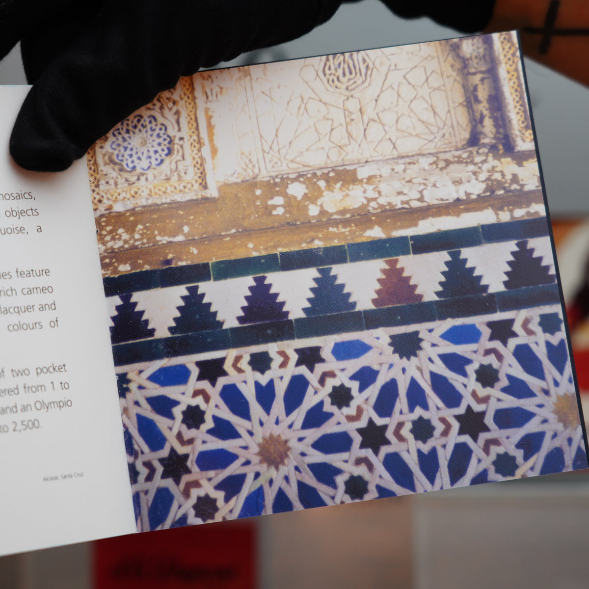 A gloved hand holds an open book displaying ornate tile patterns and text on the left page. The right page reveals intricate geometric designs in blue, white, and brown, reminiscent of the Vintage 2003 St Dupont Andalusia Limited Edition Blue Lacquer Table Lighter by S.T. Dupont, one of only 300 made.