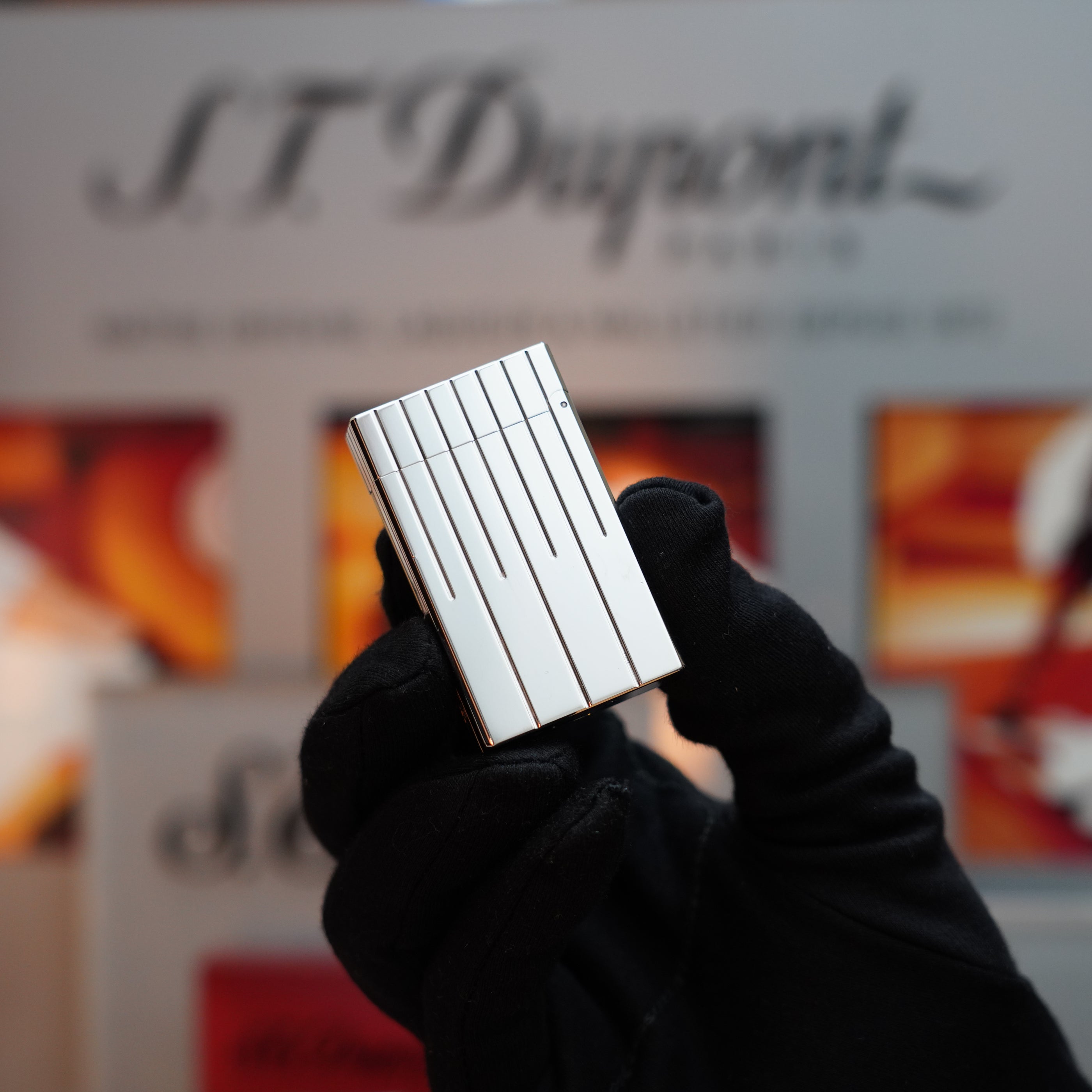 A gloved hand holds a Vintage 1999 St Dupont Rare Perspective 2000 Palladium Limited Edition Ligne 1 Lighter with vertical grooves against a blurred background displaying the S.T. Dupont logo.