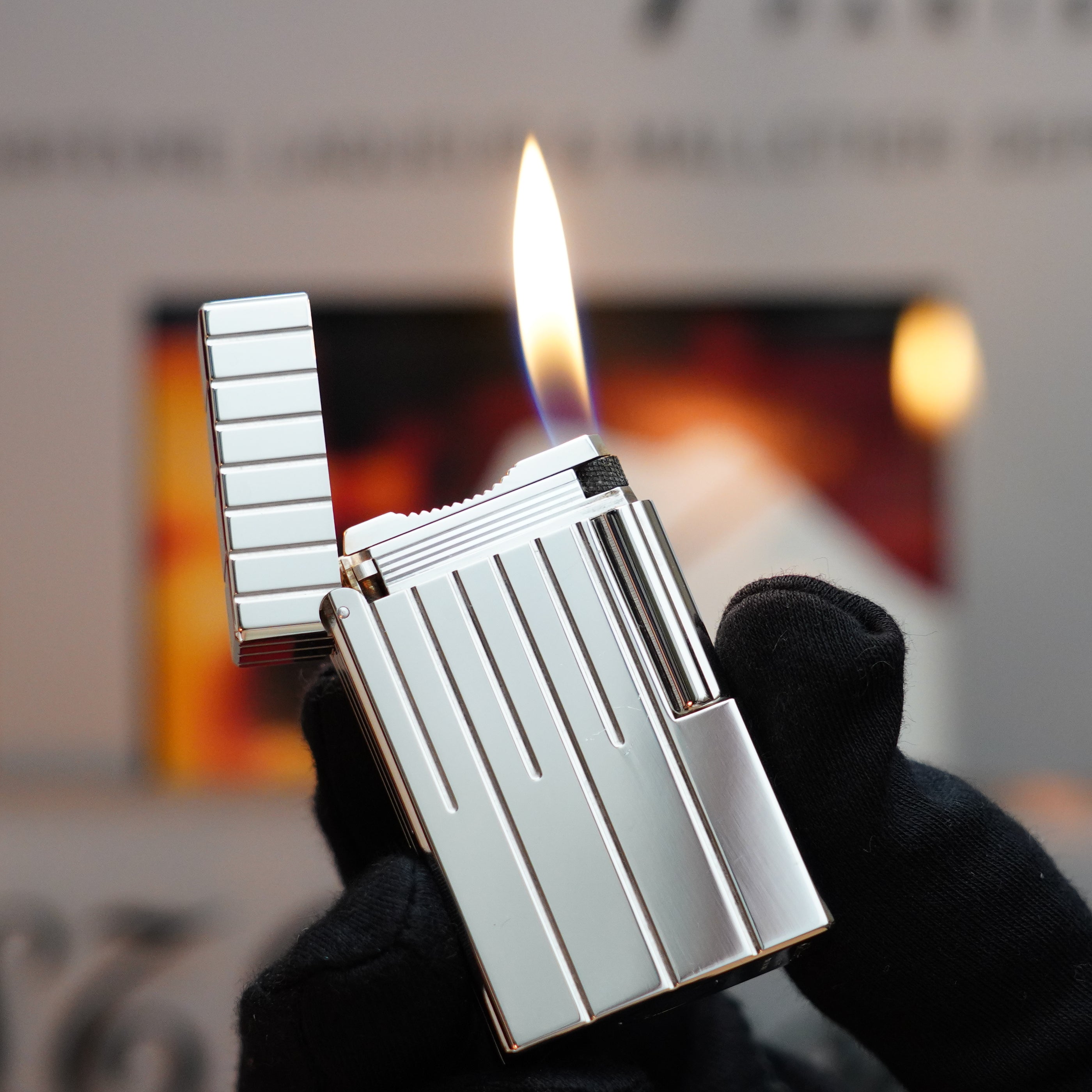 A hand in a black glove holds an ignited Vintage 1999 S.T. Dupont Rare Perspective 2000 Palladium Limited Edition Ligne 1 Lighter, featuring a vertical striped design, against a blurred background.