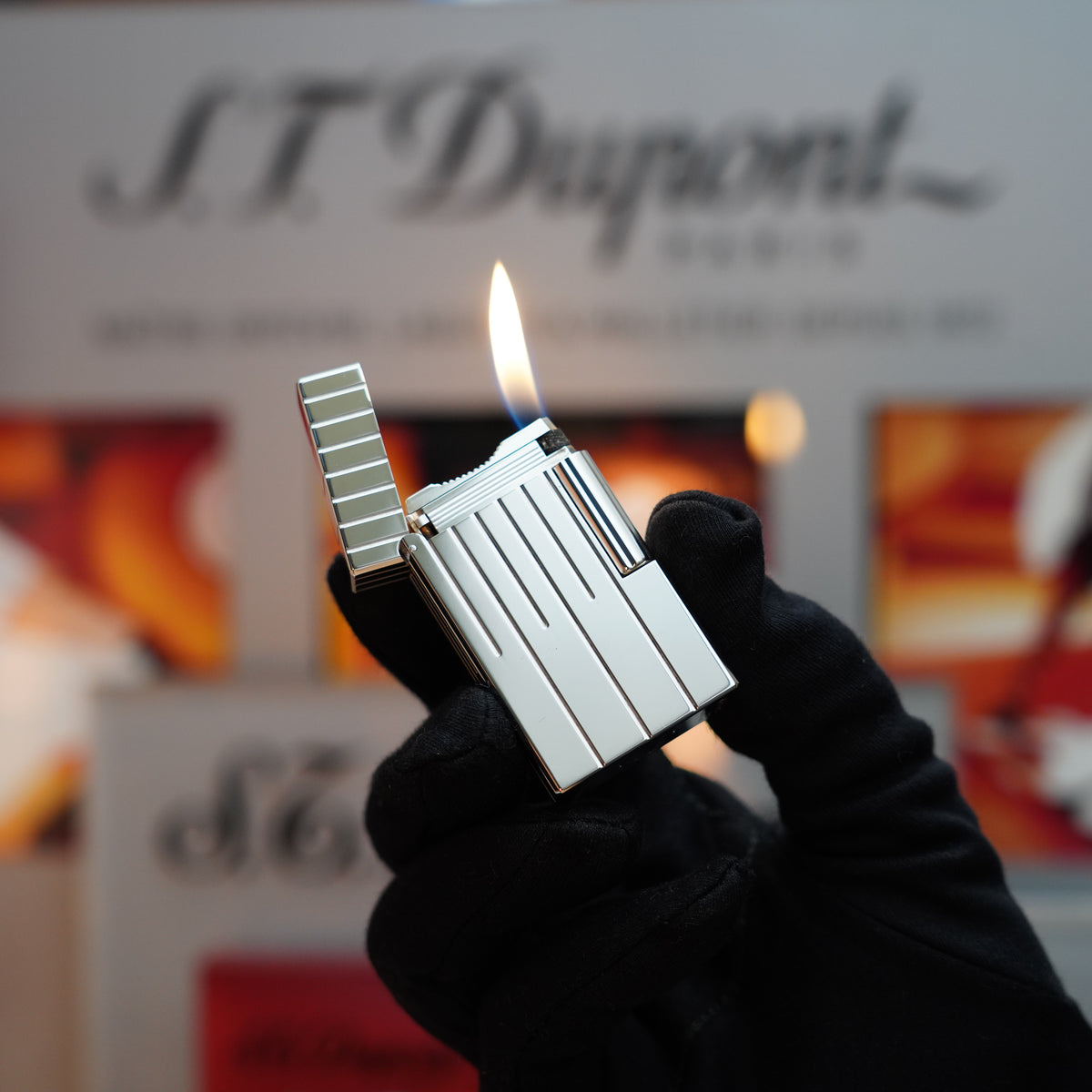 A gloved hand holds a silver flame from the Vintage 1999 St Dupont Rare Perspective 2000 Palladium Limited Edition Ligne 1 Lighter, against a blurred backdrop showcasing the text "S.T. Dupont.