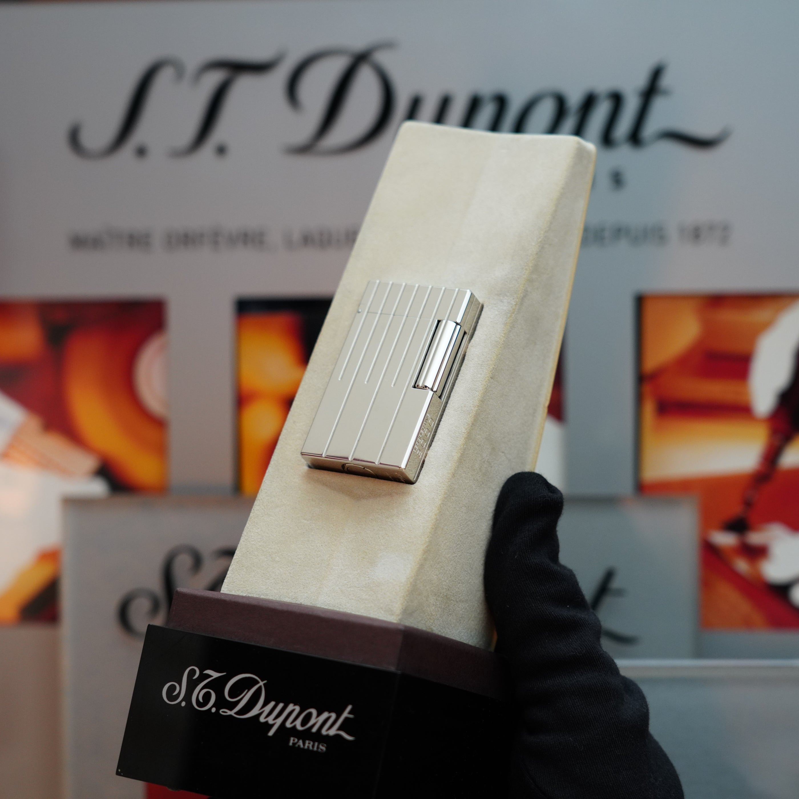 A person wearing a black glove holds the Vintage 1999 St Dupont Rare Perspective 2000 Palladium Limited Edition Ligne 1 lighter in front of a display featuring the S.T. Dupont brand, highlighting its timeless elegance.