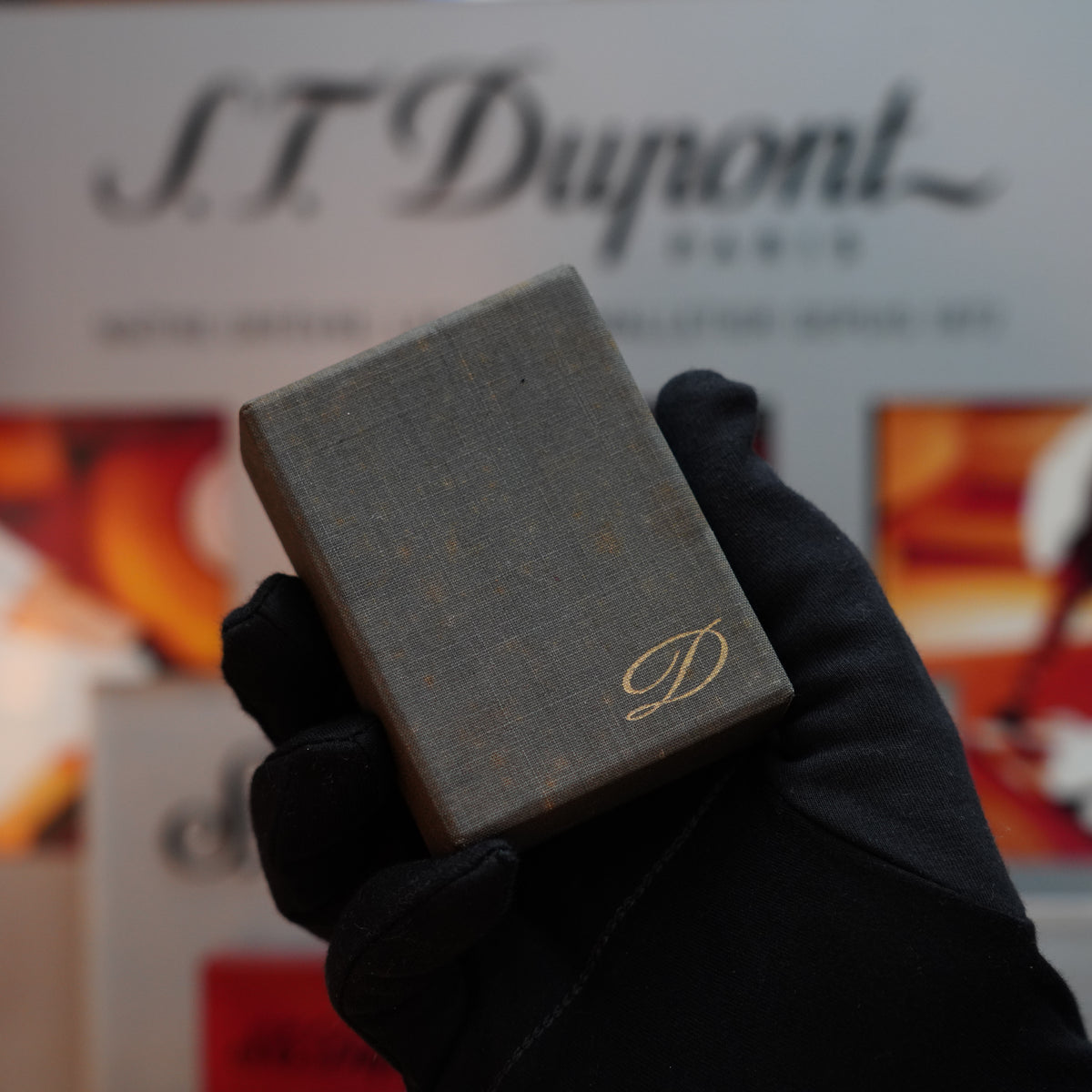 A gloved hand holds a small, textured box featuring the iconic gold "D" on the front, reminiscent of an S.T. Dupont creation. The background is softly blurred with text and images that evoke luxurious vibes, akin to a Vintage 1970 S.T. Dupont Ligne 1 Lighter with its rare visual pattern and opulent 24k gold finish.
