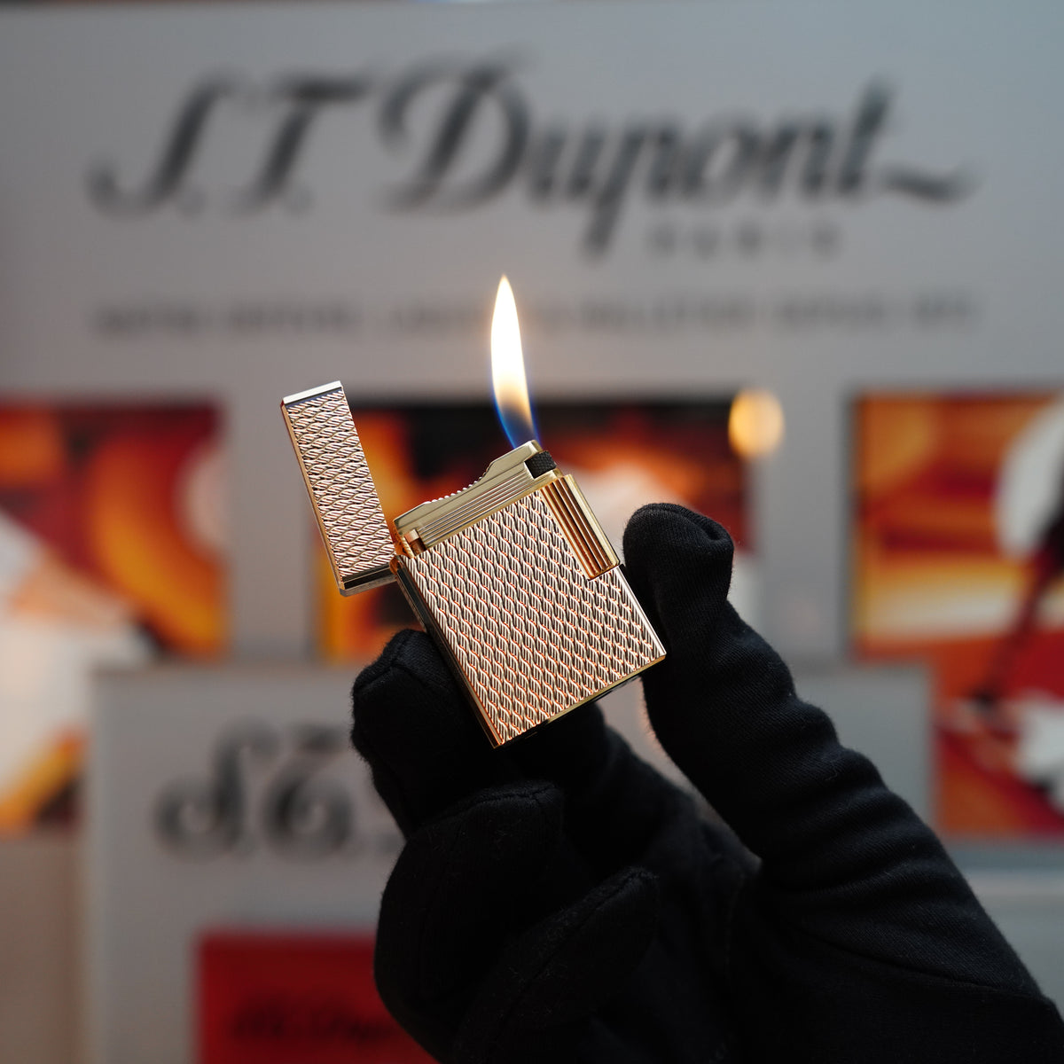 A gloved hand holds an open S.T. Dupont Vintage 1970 Ligne 1 lighter, showcasing its heavy 24k gold finish with a rare visual pattern, in front of a blurred background featuring text and graphics.
