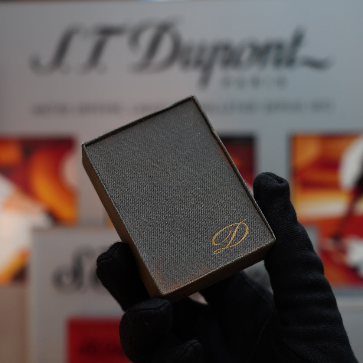 A gloved hand holds a small box emblazoned with a gold "D", unveiling the rare collectible inside—a Vintage 1970 S.T. Dupont Ligne 1 lighter featuring a stunning heavy 24k gold finish and rare visual pattern—set against a blurred background showcasing the brand's name.