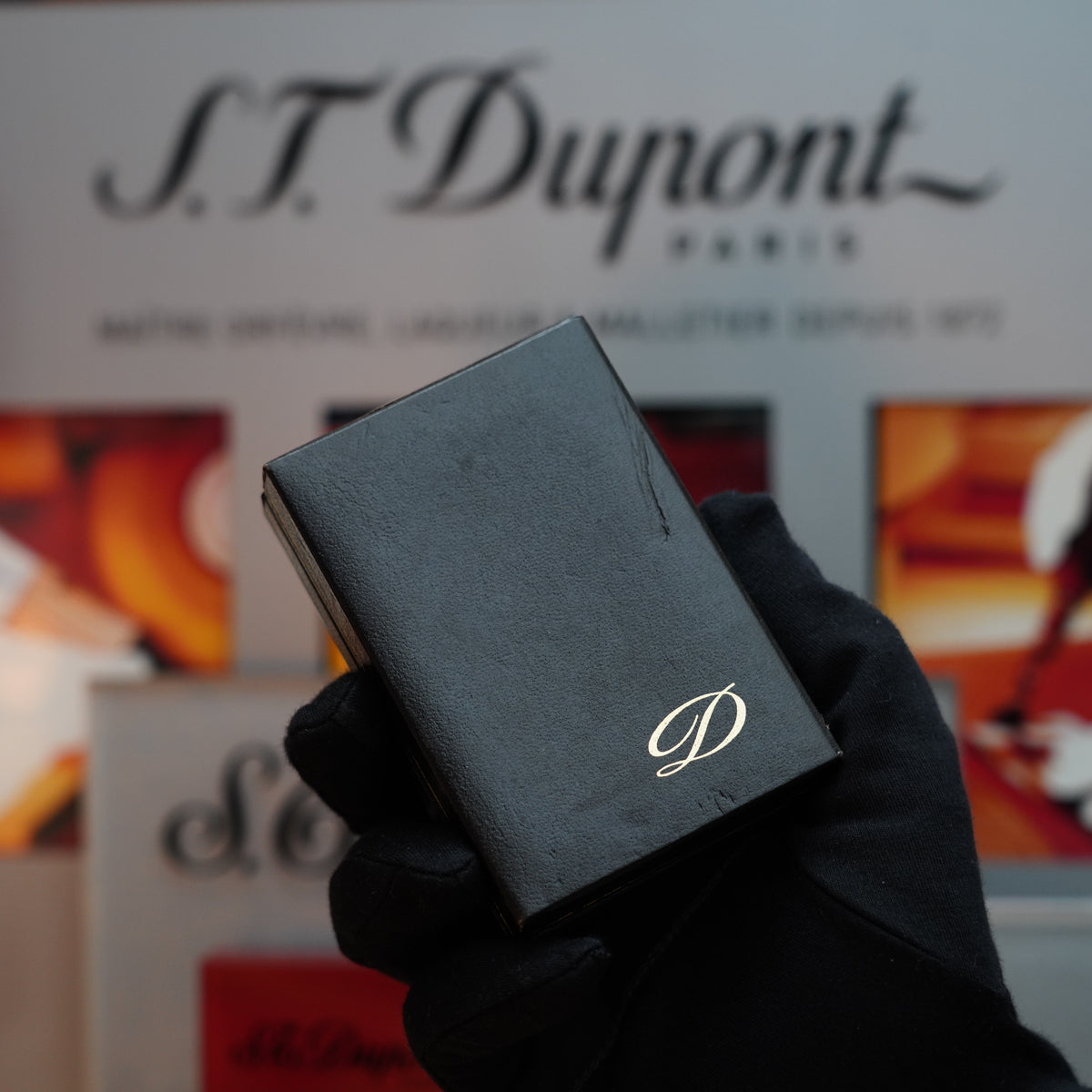 A gloved hand holds a black box adorned with a "D" emblem, suggesting the luxury of a 1988 S.T. Dupont Gatsby Gainerie Lezard Brown lighter with a 24k gold finish and authentic leather, positioned in front of an S.T. Dupont display.