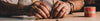 Hands of a person measuring fabric with a tape measure on a table, with a red and beige striped container nearby.