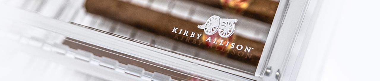 Close-up of a transparent case with a visible logo that reads "Kirby Allison" and contains several cigars inside.
