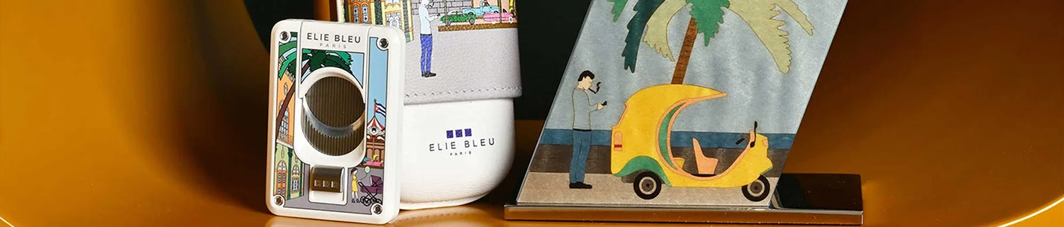 A colorful cigar cutter, a white case, and a decorative card holder with a tropical car illustration are displayed on a gold surface.