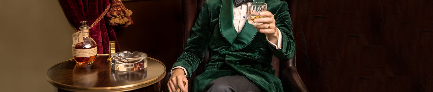 A person in a green velvet suit sits in a chair, holding a glass of whiskey. A round table nearby has a bottle of whiskey and a glass ashtray. The scene suggests a formal, elegant setting.