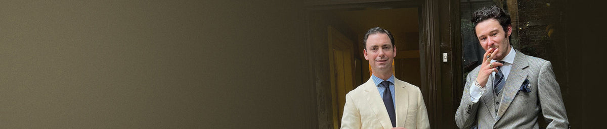 Two men in tailored suits standing in front of a doorway, one smoking a cigar.