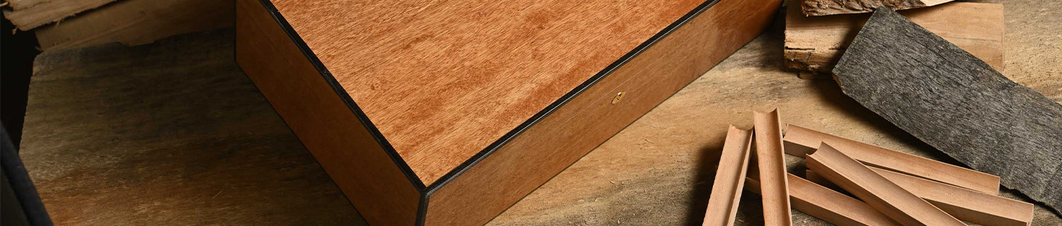 A wooden box sits on a wooden surface next to several thin wood strips and textured materials.