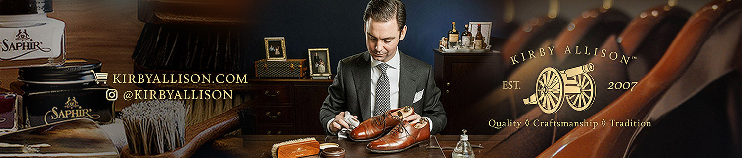 A man polishing brown leather shoes at a wooden desk with shoe care products, branding for Kirby Allison, established 2007, displayed in the background.