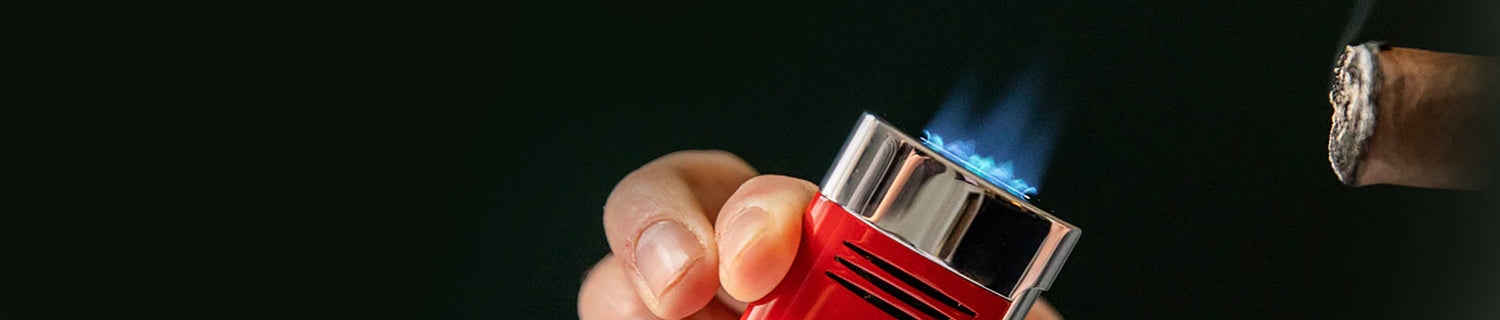 A hand holds a red torch lighter with a blue flame, aimed at the end of a cigar against a dark background.