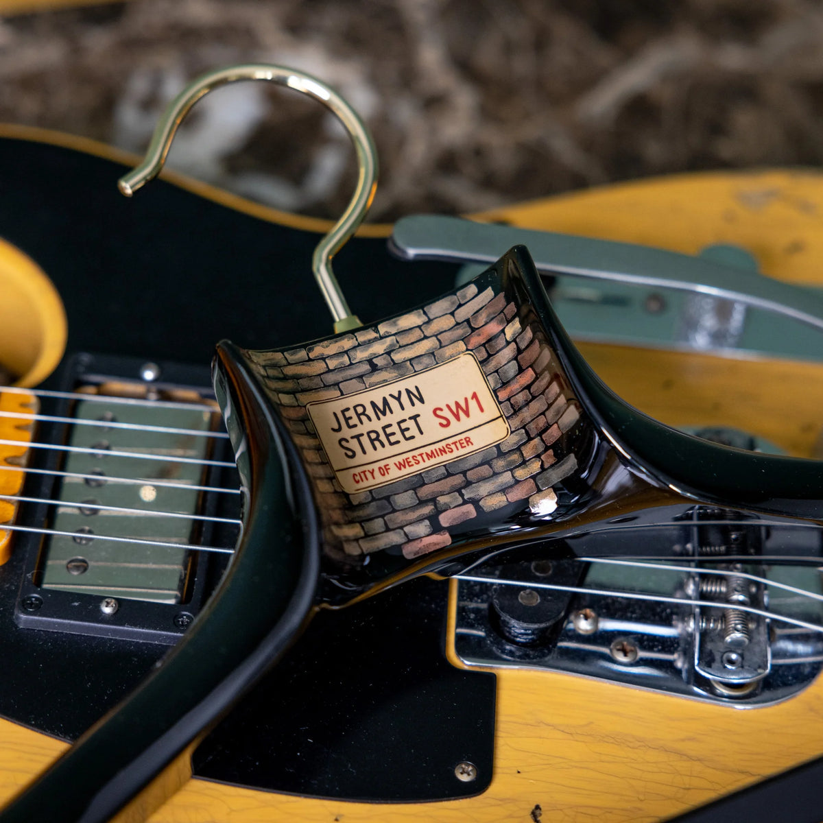 A Nakata Wajima Urushi Jermyn Street Limited Edition Hanger from KirbyAllison.com, featuring a street sign that reads "Jermyn Street SW1, City of Westminster," is resting on the strings of a black and yellow electric guitar, next to a finely crafted piece of hand-planed wood.