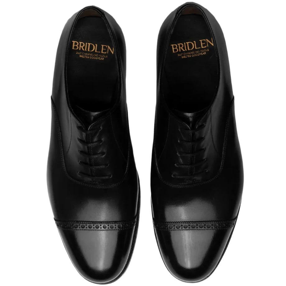 A pair of black Bridlen Punch Cap Oxford Rui Last dress shoes in box calf leather with decorative stitching, perfect for formal occasions, elegantly displays the brand name on the insoles.