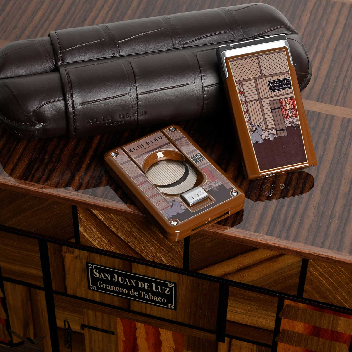 A close-up styled product shot showing a cigar humidor and matching cigar case on a wooden surface, with a branded cigar box in the background. The humidor has a brown leather-like finish and printed illustration panels, and the case is metallic silver with a matching printed panel.