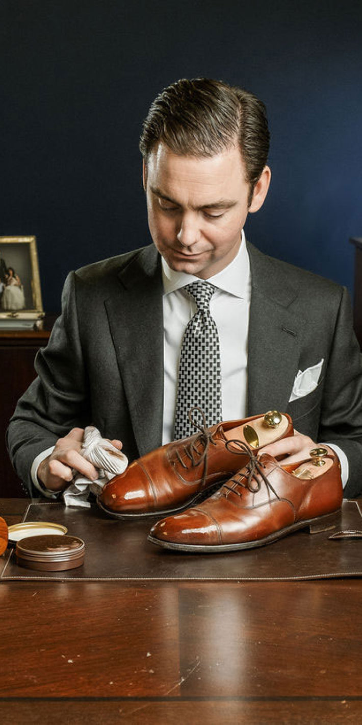 Image of Kirby Allison seated at a desk, wearing a suit, and tie and looking down, while polishing a pair of brown dress shoes. 